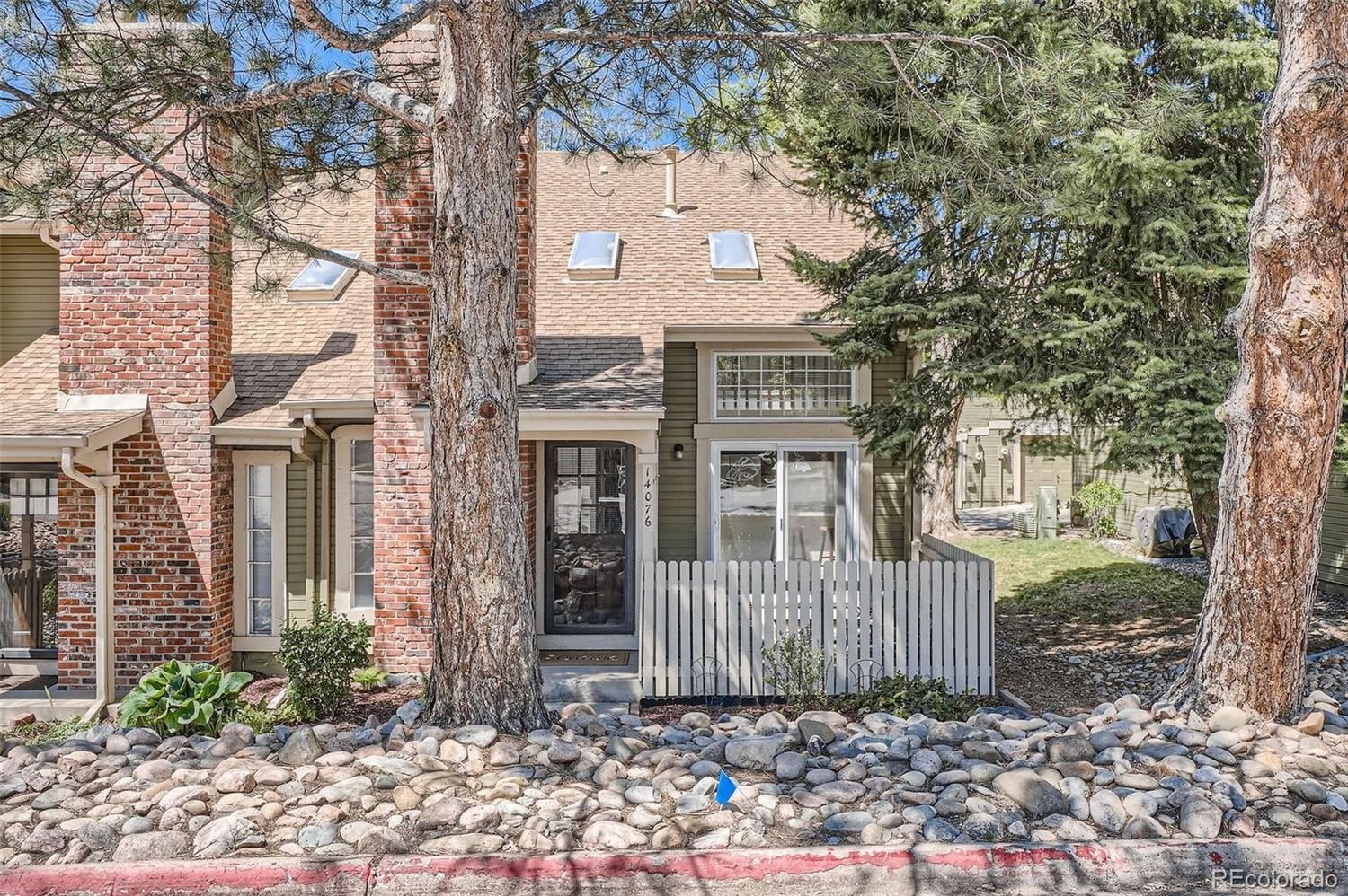 Front view of a townhouse with brick and green siding, a small enclosed porch, large pine trees, and rocky landscaping.