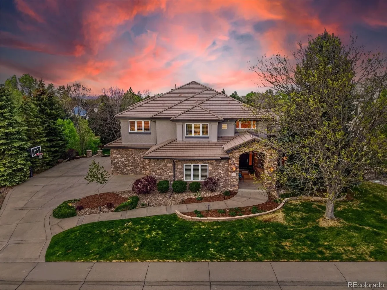 A large two-story house with a stone and gray exterior, surrounded by trees and a well-maintained lawn, during a colorful sunset with pink and purple clouds.