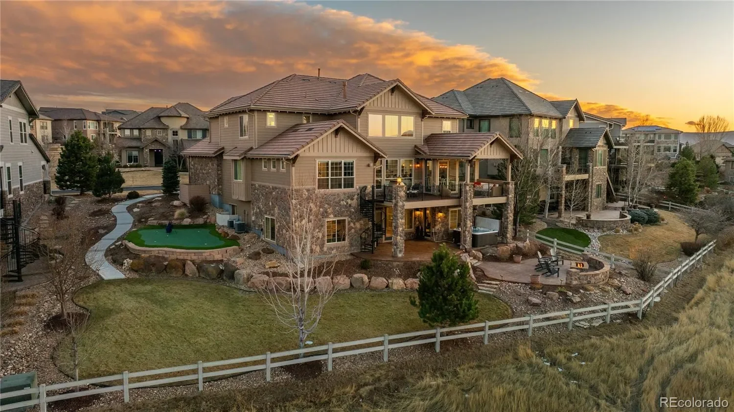 Large multi-story suburban house with stone and siding exterior, surrounded by landscaped yard with trees, pathways, and a small putting green, at sunset.