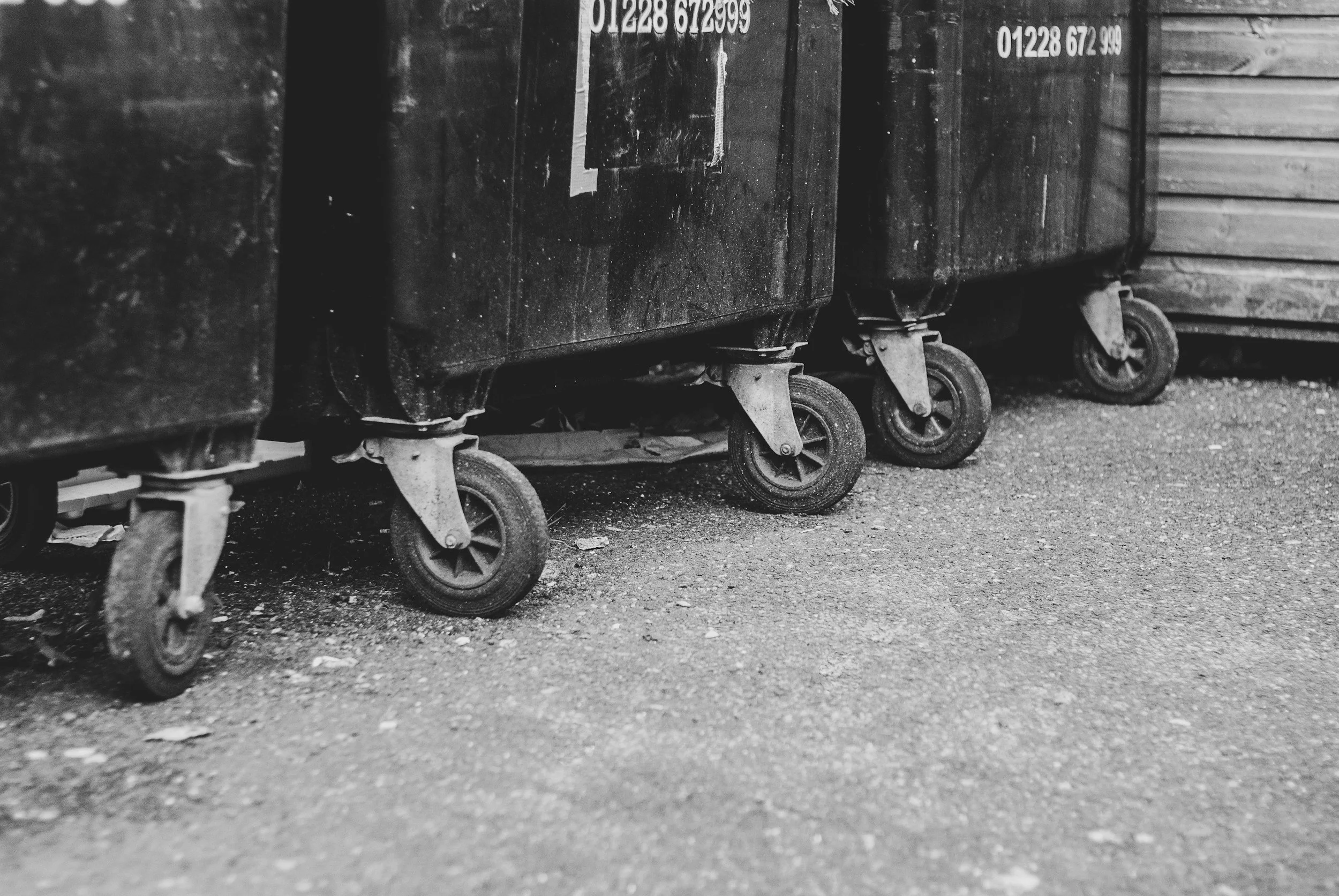 A row of black trash bins on wheels lined up outside on a gravel surface, with some stickers displaying phone numbers on their sides.