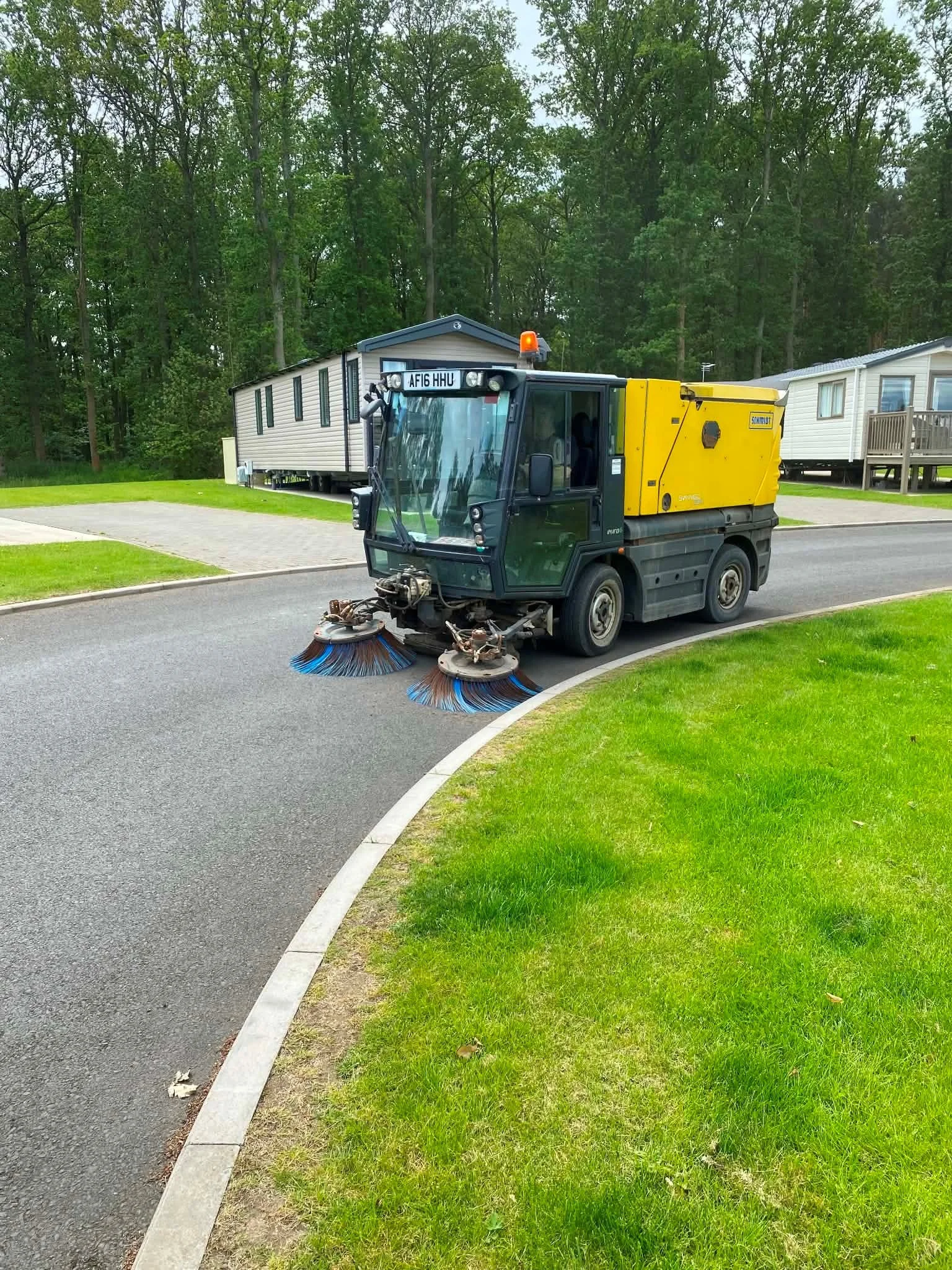 A street sweeper vehicle cleaning a paved road in a residential area with mobile homes and green trees in the background.