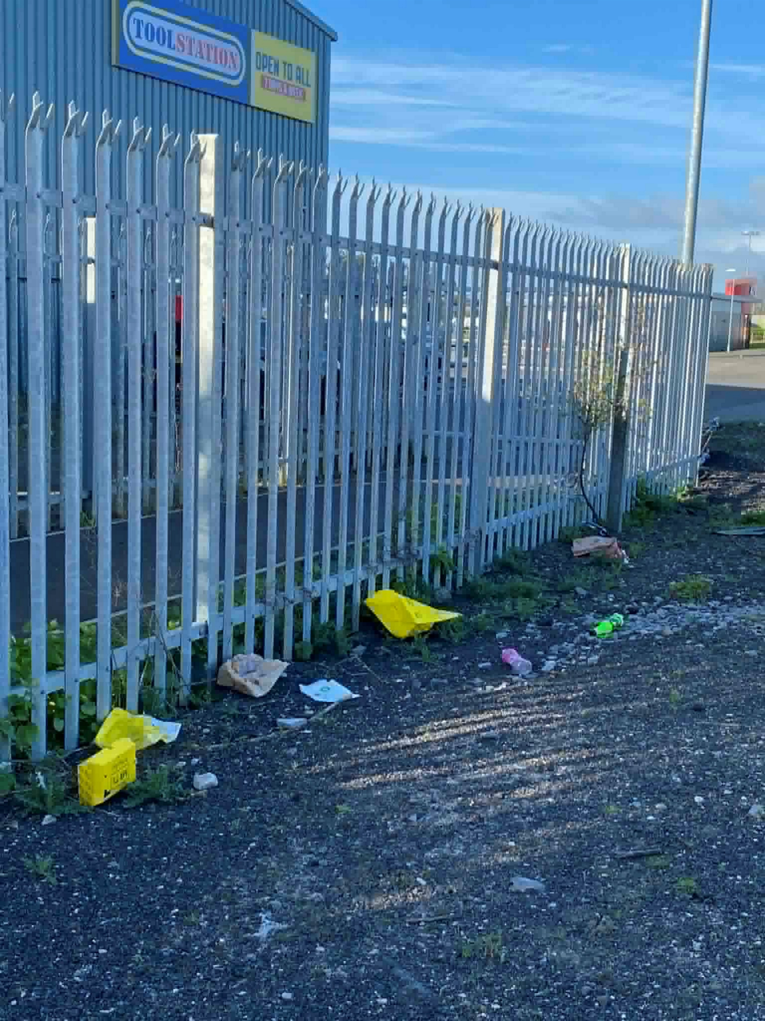 A chain-link fence beside a blue building with a sign that reads 'Tool Station' and 'Open to All.' The ground in front of the fence is littered with trash, including yellow and pink plastic items, paper, and other debris, with a clear blue sky overhead.