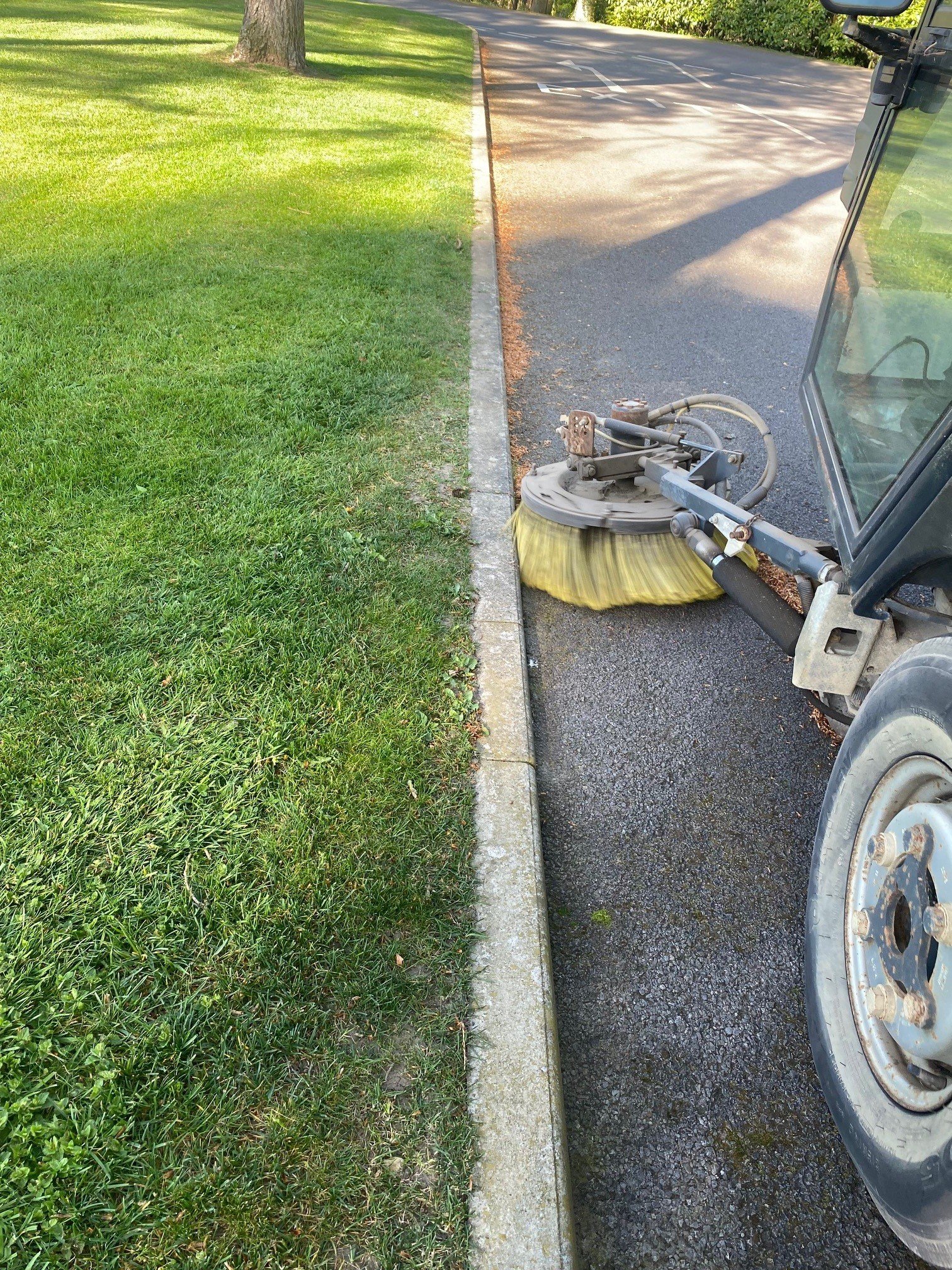 A street sweeper machine cleaning the edge of a paved road next to a grassy area with trees.