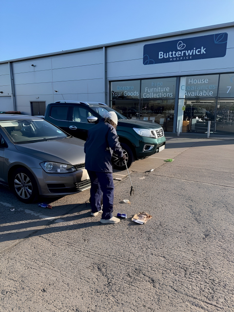 Person cleaning up trash in parking lot outside Butterwick Hospice, near parked cars, with store windows in background.