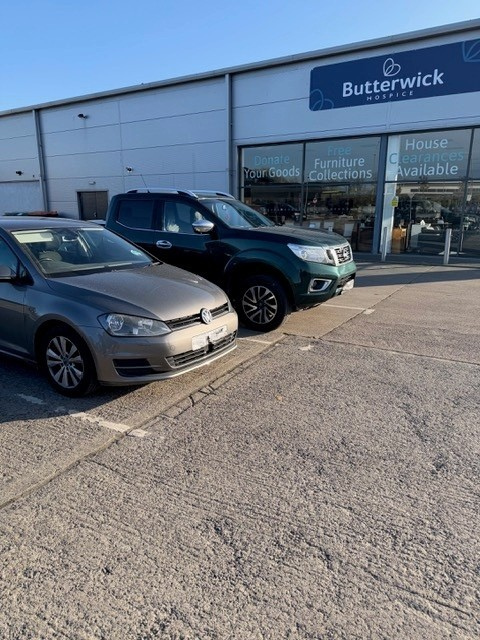 Car parked outside Butterwick Hospice store, which sells furniture and household items. The store has large windows with signs advertising donations, furniture collections, and house clearance services.
