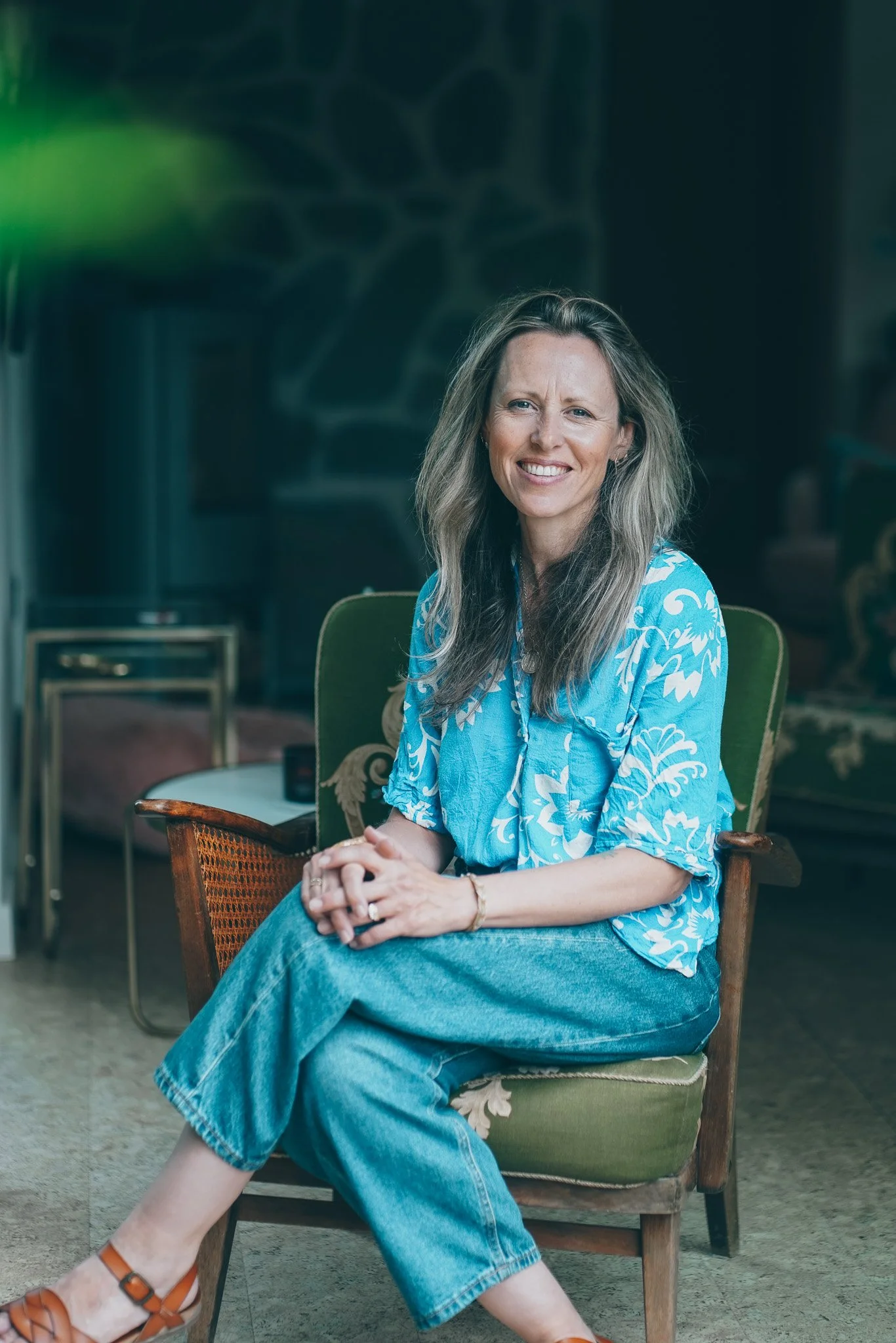 A woman with long wavy blonde hair, smiling, wearing a blue and white patterned top, jeans, and tan sandals, sitting on a green vintage chair in a cozy indoor setting with dark walls and decorative furniture.