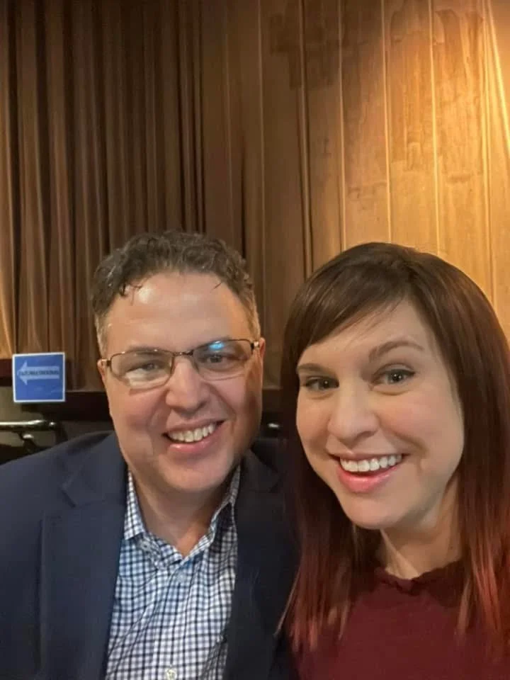 A man and woman smiling for a selfie in an indoor setting with wooden paneled walls.