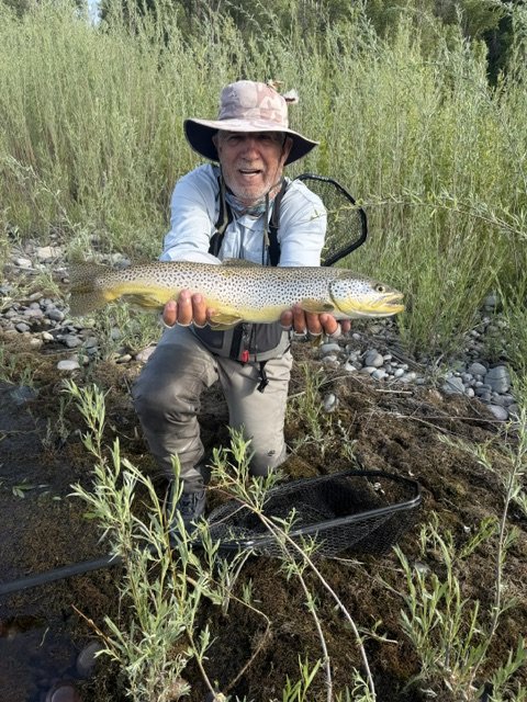 Author Mark Wierzbinski in outdoor clothing and a wide-brimmed hat kneeling in a riverbank holding a large fish with both hands, with tall grass and rocks behind him.