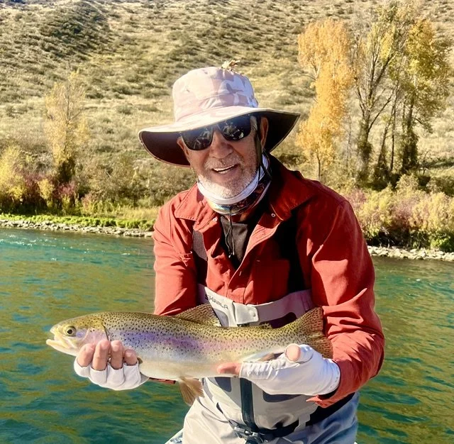 Author Mark Wierzbinski holds a large rainbow trout he caught while fishing in a river, with colorful trees and hills in the background.