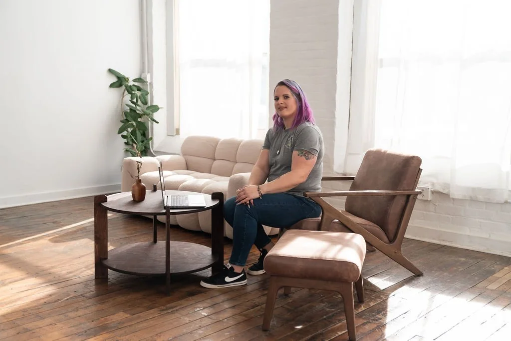 A woman with purple hair sitting on a lounge chair in a bright, modern living room with white walls, a beige sofa, wooden flooring, and large windows.