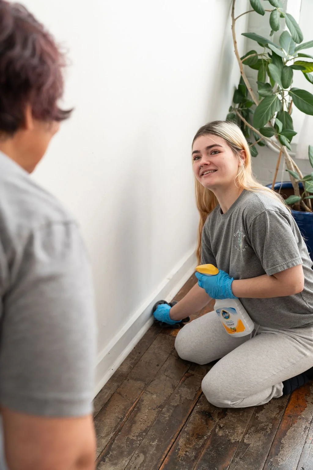 A young woman is kneeling on a wooden floor, cleaning a baseboard with a brush. She is wearing gloves and has a spray bottle attached to her pants. She is smiling and talking to an older person, who is partially visible and facing her. There is a large potted plant in the background near a window with white curtains.
