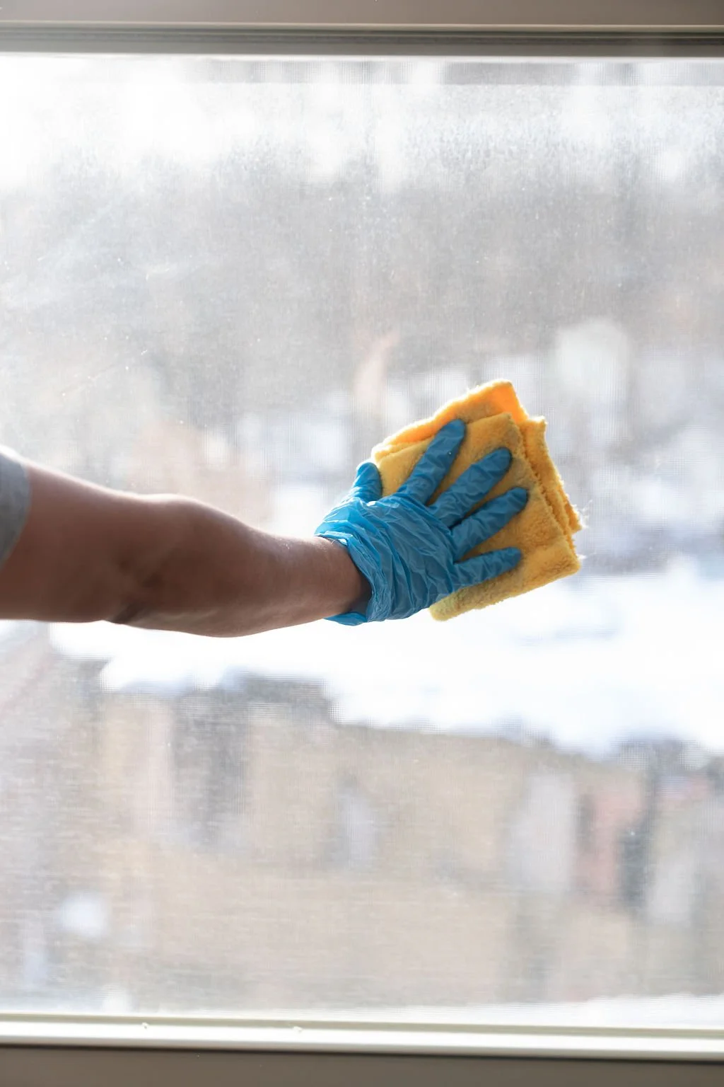 Person wearing a blue glove cleaning a large window with a yellow sponge, with snow visible outside.
