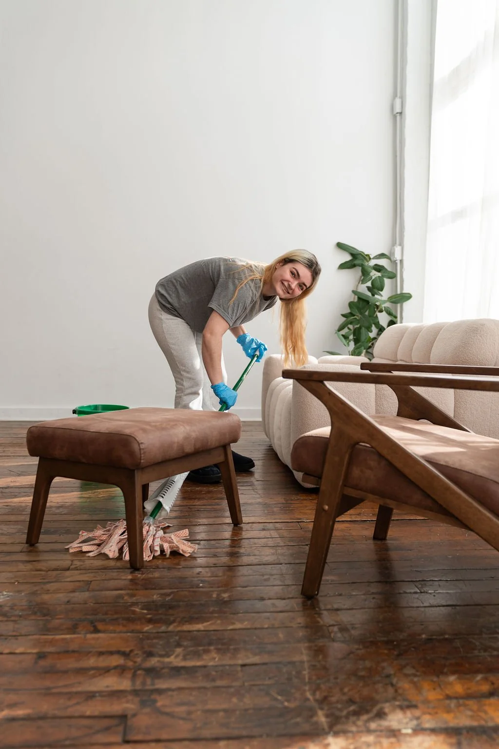 A woman is cleaning a wooden floor in a living room, using a mop with a cloth wrapped around the mop head. She is smiling and wearing a gray t-shirt, white pants, blue gloves, and black shoes. There is a beige sofa, a brown ottoman, and a wooden armchair in the room, with a large window and a green plant in the background.