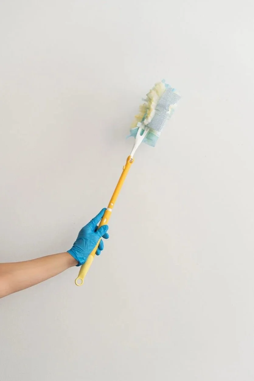 A person wearing a blue glove is holding a yellow mop with a cleaning cloth attached to the head, cleaning a plain white wall.