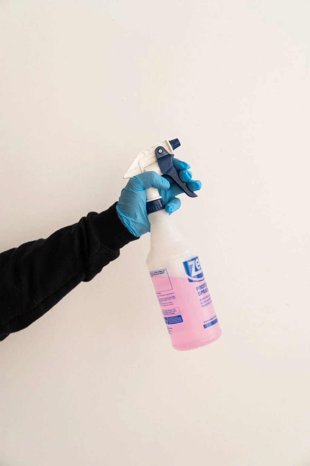 A person wearing a black long sleeve shirt and blue latex gloves holding a pink spray bottle against a plain white wall.