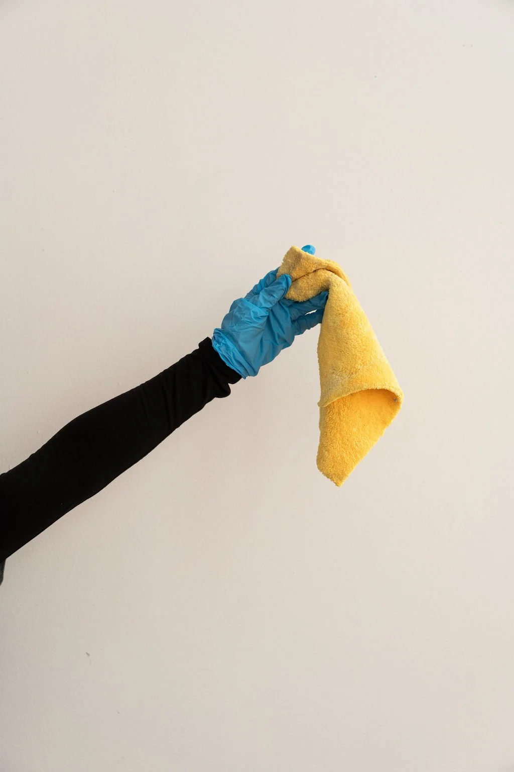 Person wearing a black long sleeve shirt and blue glove holding a yellow microfiber cloth against a plain white wall.