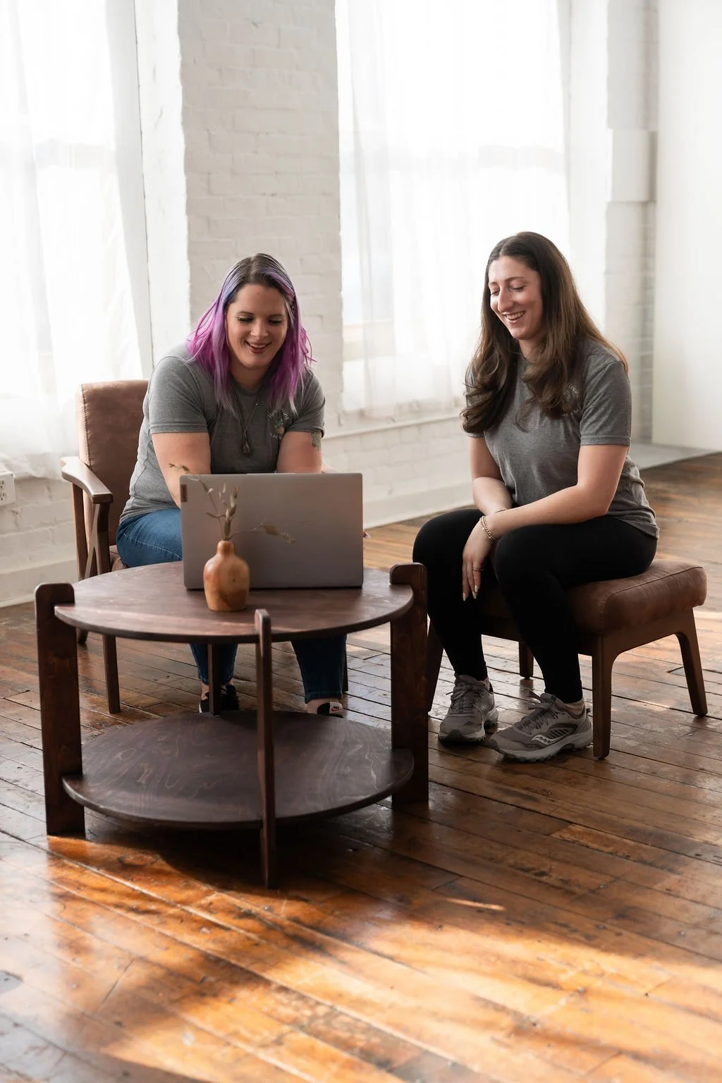 Two women sitting on chairs in a well-lit room, looking at a laptop on a wooden coffee table with a small vase and decorative branch, engaging in a conversation and smiling.