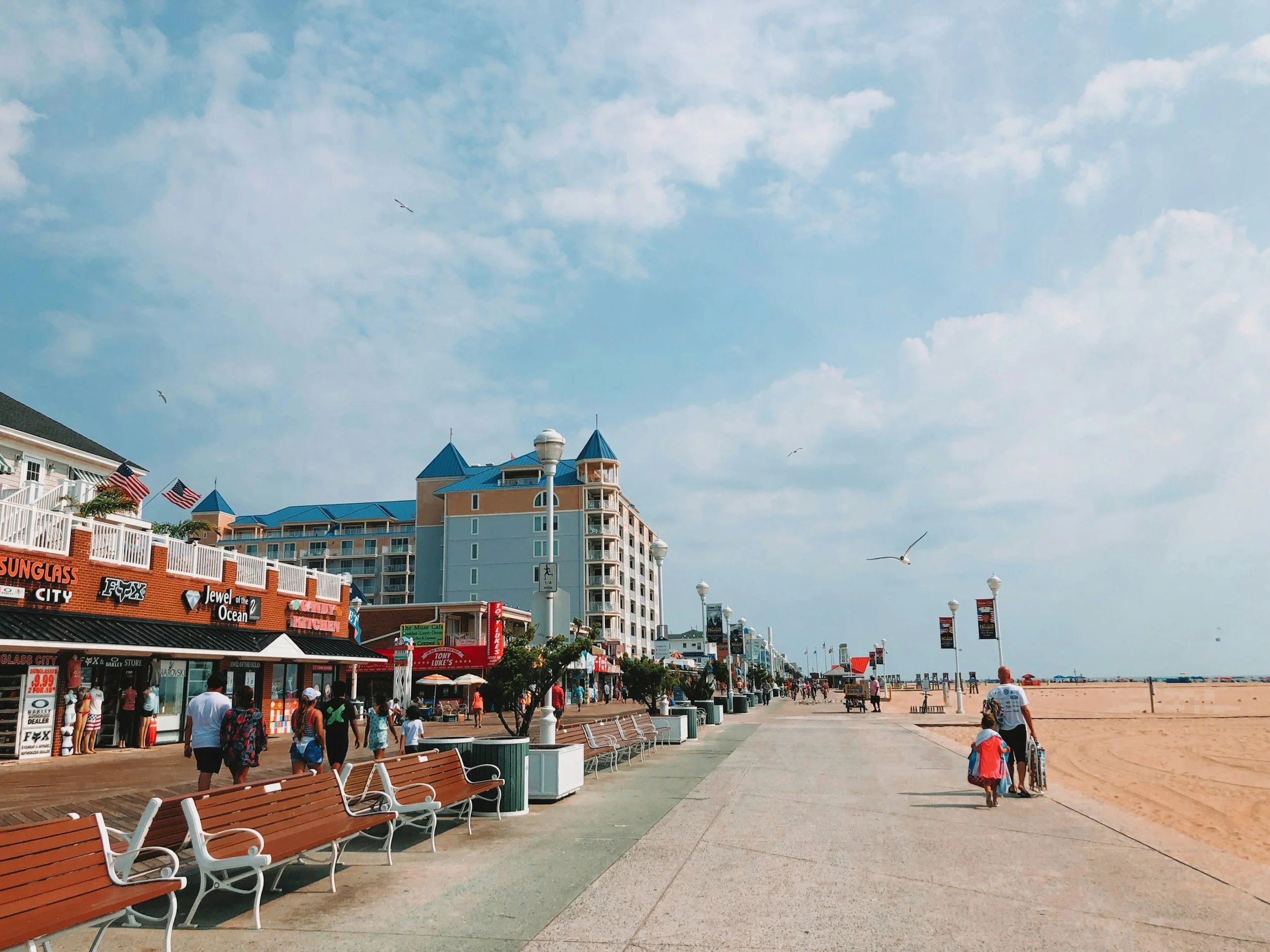 Beachside promenade with shops on the left and the ocean on the right. People walk along the path, and seagulls fly in the sky. There are benches, lampposts, and flags visible.