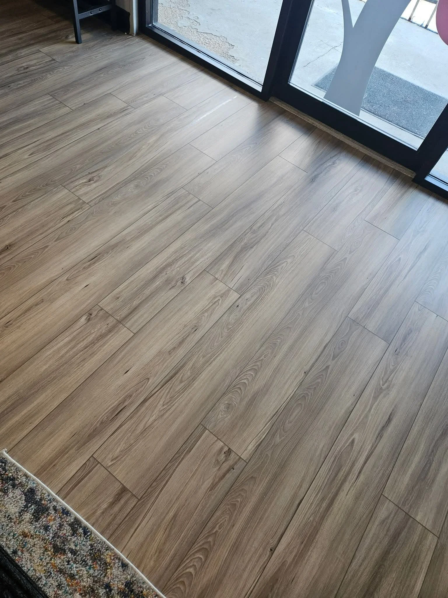 View of a hardwood floor near a glass door leading outside, with part of a patterned rug visible in the bottom left corner.