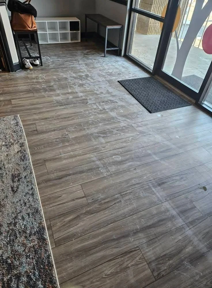 Interior space with wood-look flooring, a black doormat, glass door, and minimal furniture including a small table, a bench, and a white storage shelf.