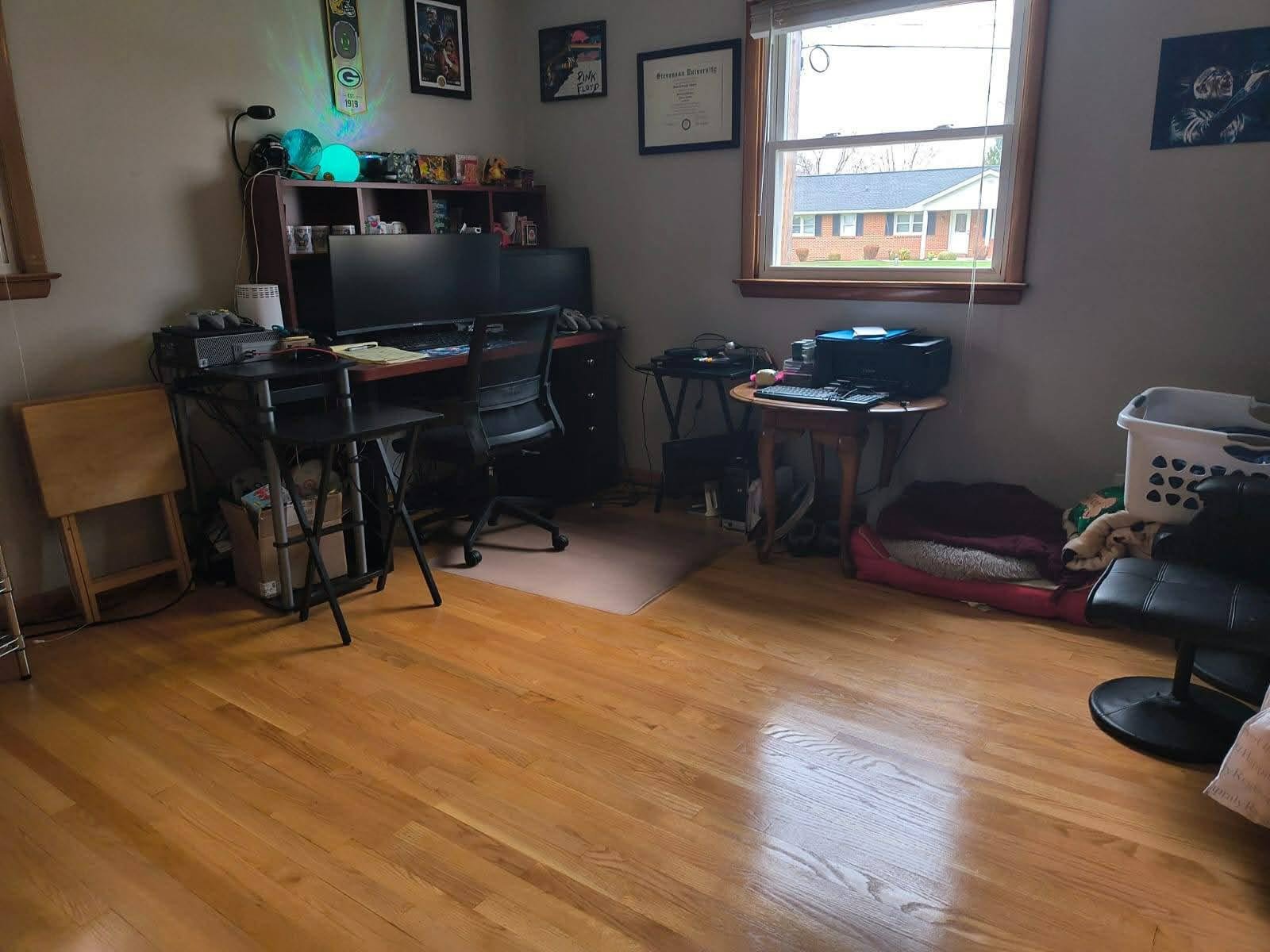 A room with wooden floors and a large window. There is a black desk with multiple monitors, a chair, and various electronic devices. To the right, there's a small table with a printer and other items. On the floor, there are pet beds, blankets, and a laundry basket. The walls have framed pictures and certificates.