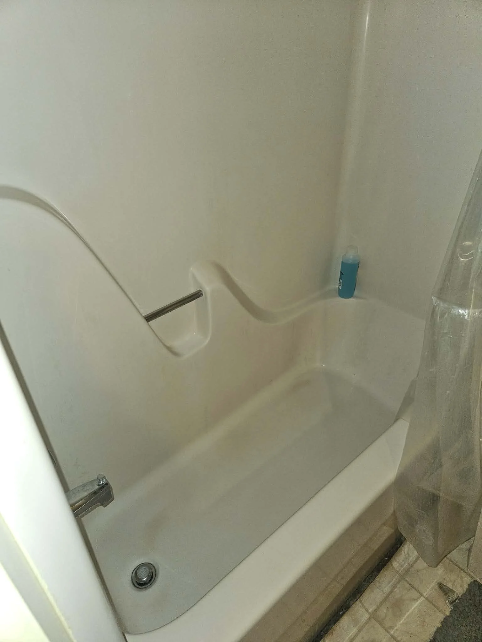 A white washing machine seen from the top, with a blue bottle of laundry detergent on top. The washing machine has control knobs and is positioned between a beige wall and a tiled floor.