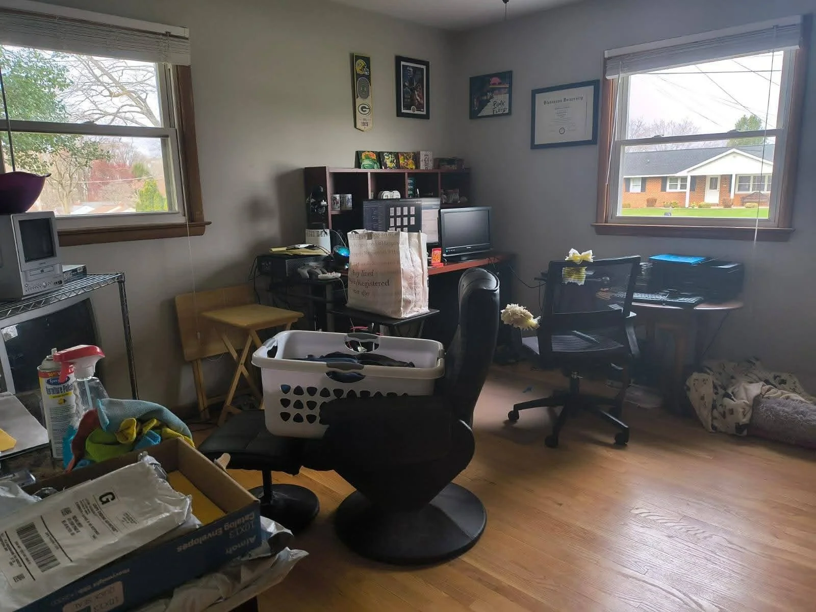 A cluttered home office with two windows, a desk with a computer and various items, a chair, a laundry basket, and framed pictures on the wall.