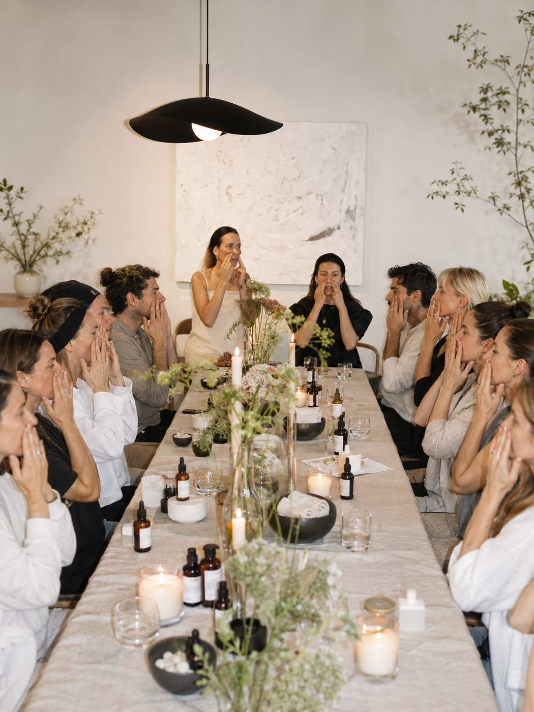 A group of people participating in a meditation session around a long dining table decorated with candles, flowers, and bottles, in a minimalist room with neutral colors and simple decor.