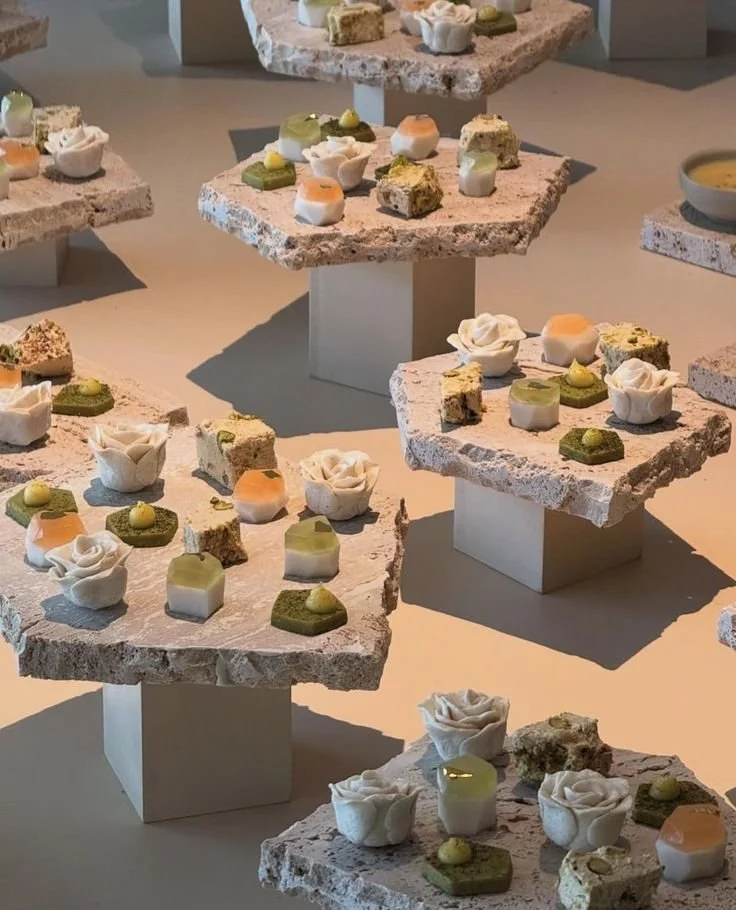 Several flat, rough-edged pink stone slabs on white stands display an assortment of small, elegantly decorated desserts including white rose-shaped confections, green and orange-topped treats, and small cake pieces in a modern setup with soft lighting.