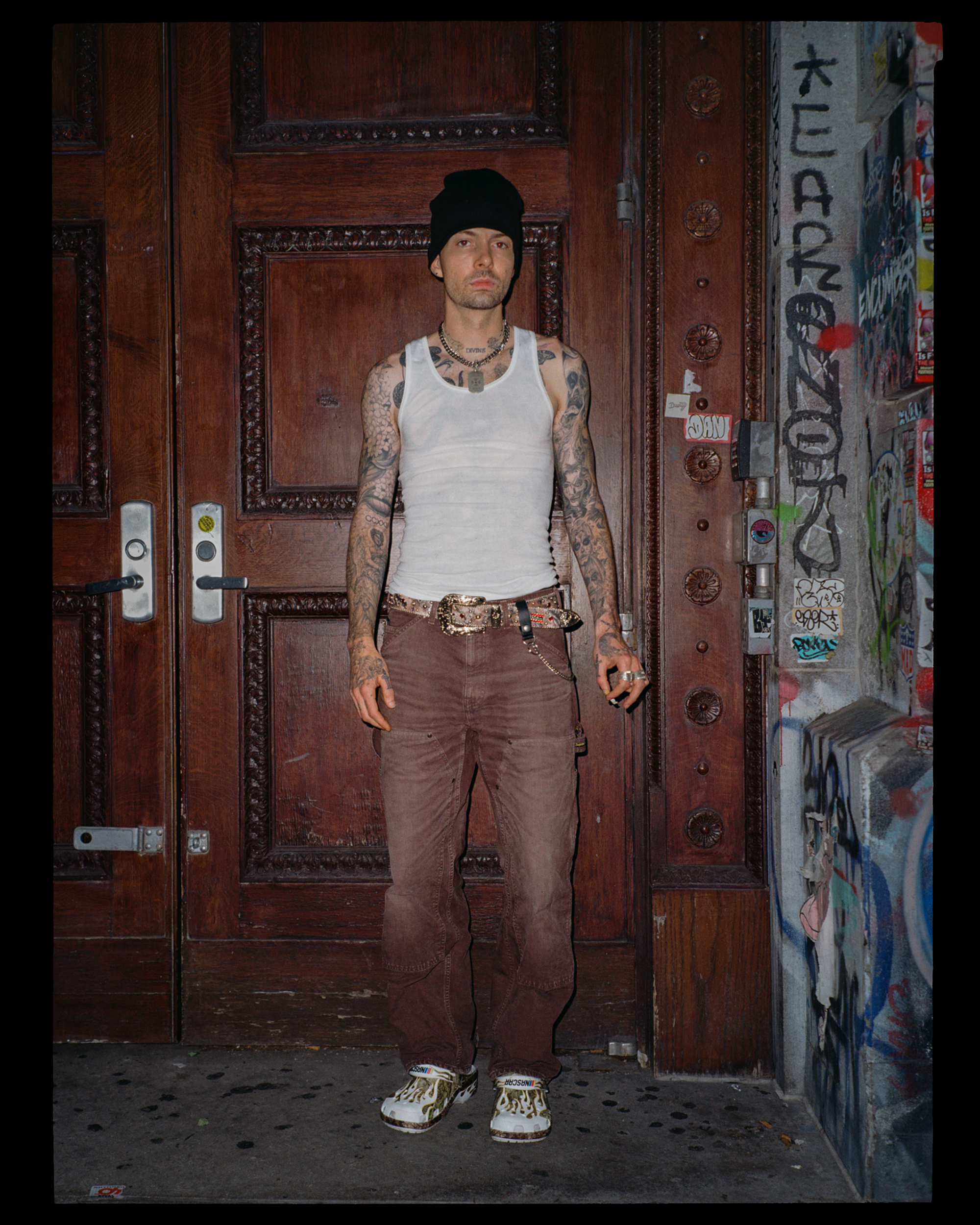 A man standing in front of a decorated wooden door with graffiti on the wall to his right, wearing a black beanie, white tank top, brown pants, and patterned sneakers, with tattoos on his arms and chest.