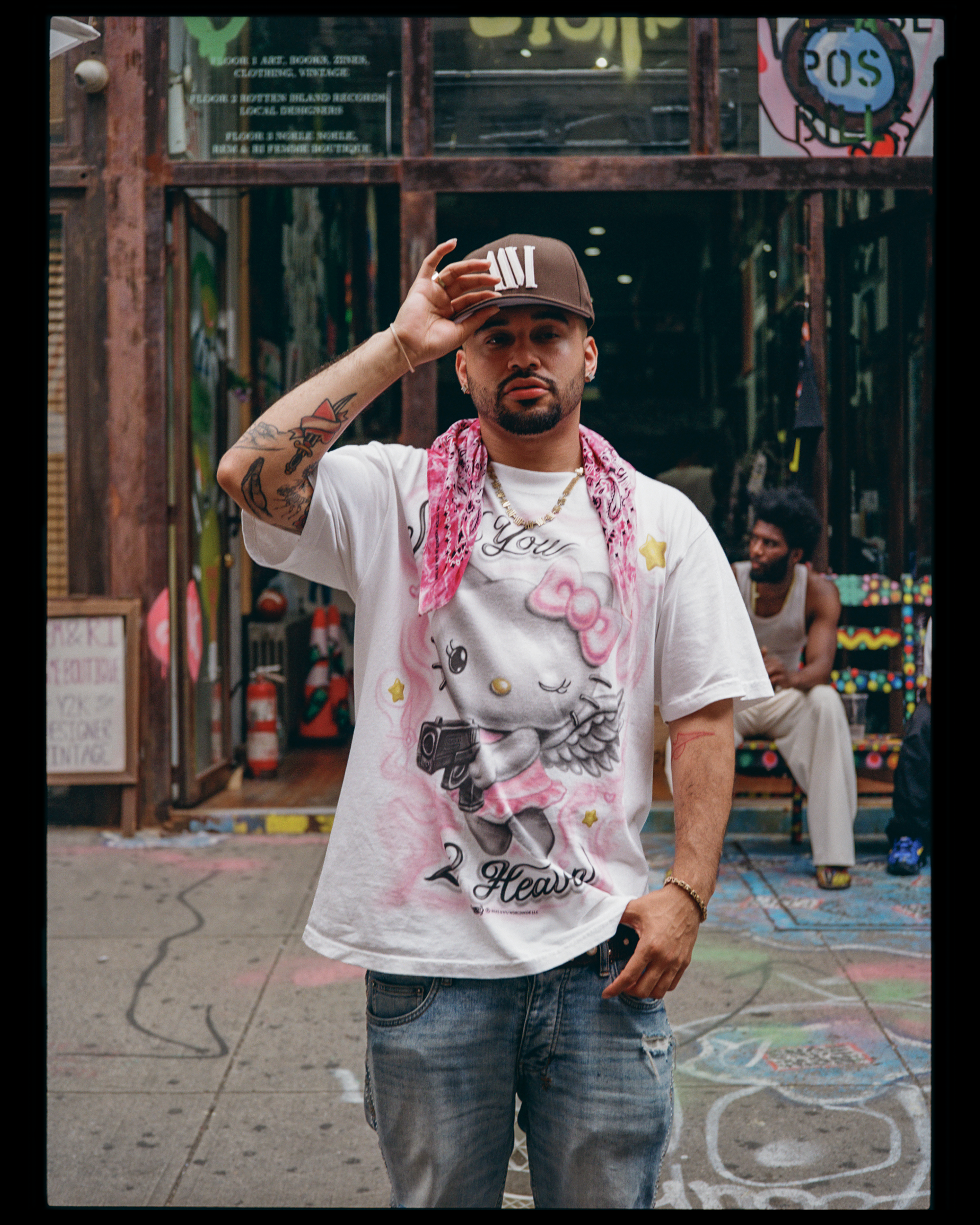 A man with tattoos on his arm and a beard standing outside, wearing a brown cap, white t-shirt with Hello Kitty design, and ripped jeans, touching his cap. In the background, there is a colorful, urban outdoor setting with a woman sitting at a table,