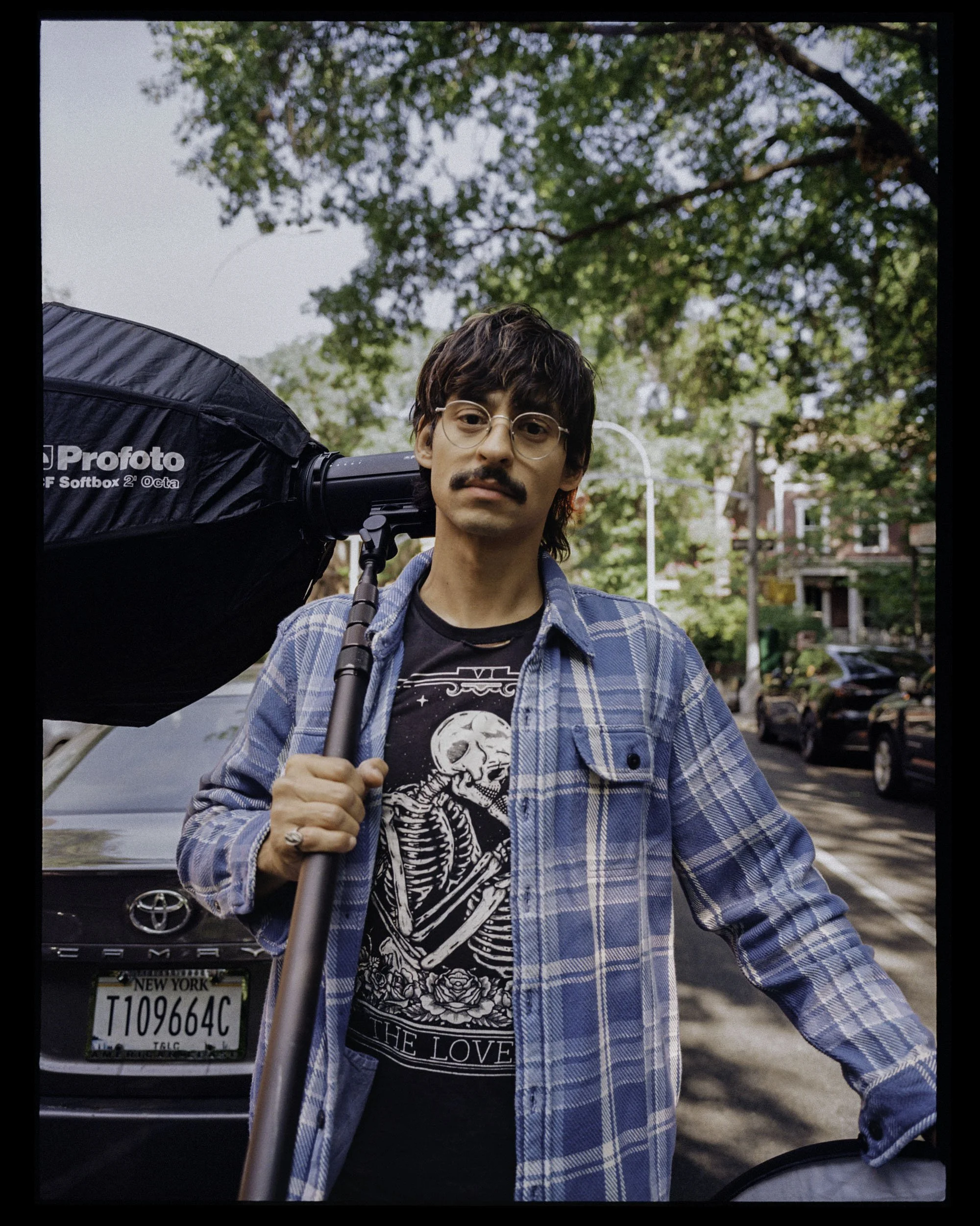 A young man with dark wavy hair, a mustache, and glasses stands outdoors, holding a large black photography softbox over his shoulder, with a background of green trees and parked cars.
