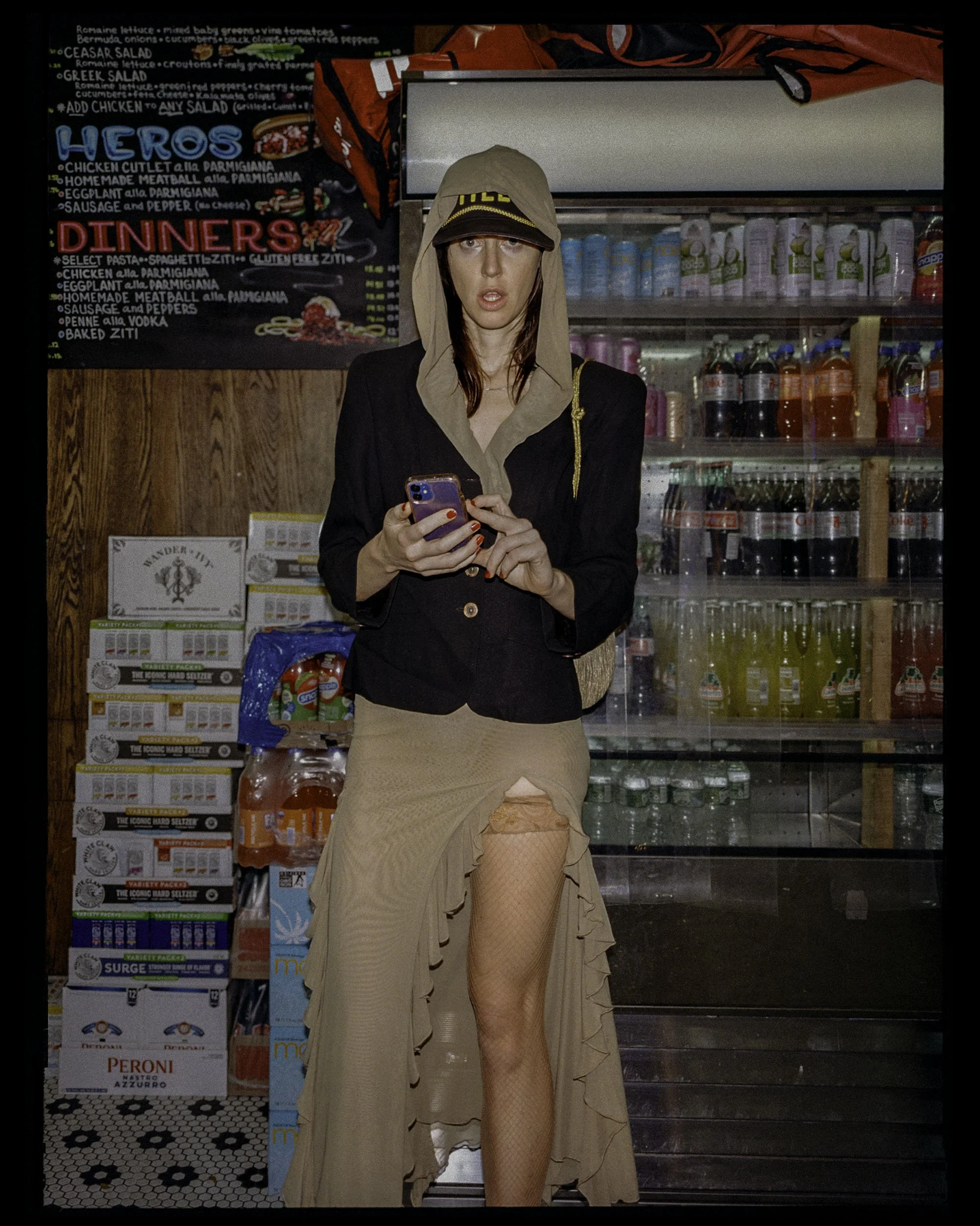 A person dressed in unconventional clothing, including a beige skirt with a high slit, fishnet stockings, and a beige hoodie with a hat, holding a phone inside a convenience store or supermarket. The background shows beverage bottles and a chalkboard