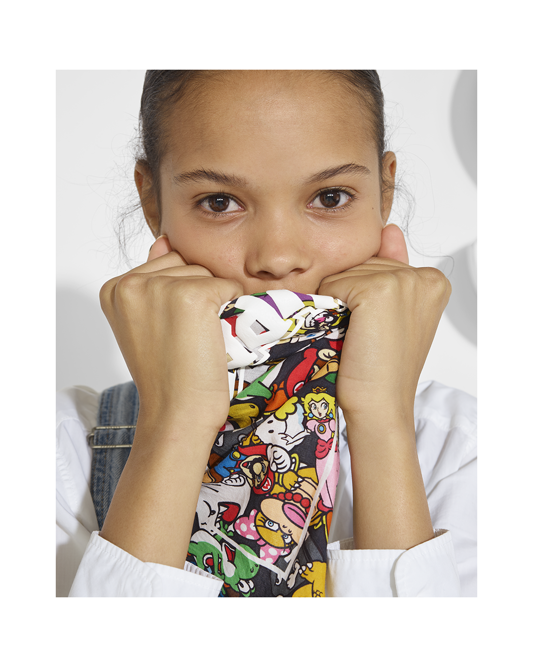 A young girl holding a colorful, cartoon-themed scarf over her mouth while looking directly at the camera.
