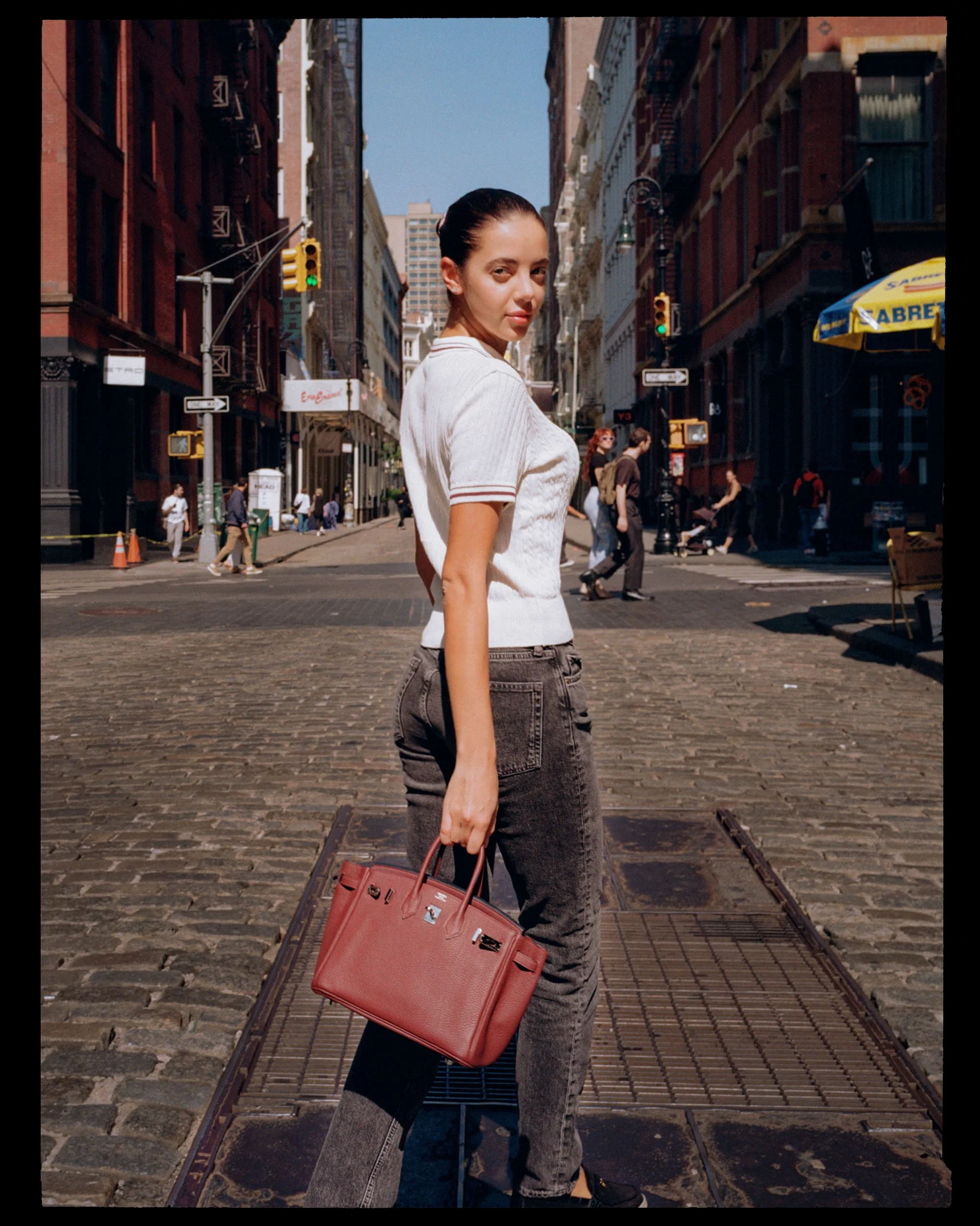 A woman with dark hair pulled back, wearing a white shirt and black jeans, standing on a city street holding a red handbag, with tall buildings, traffic lights, and pedestrians in the background.