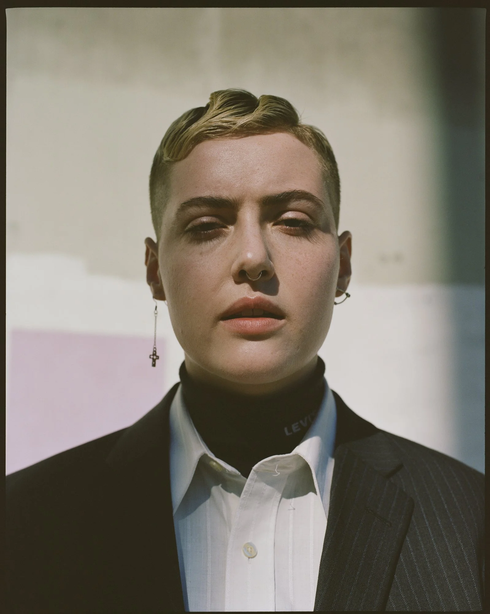 Close-up of a person with short light hair, wearing a dark pinstripe suit, white dress shirt, and a black turtleneck with the word 'Levi's' visible on the collar, with a cross earring visible on the left ear.