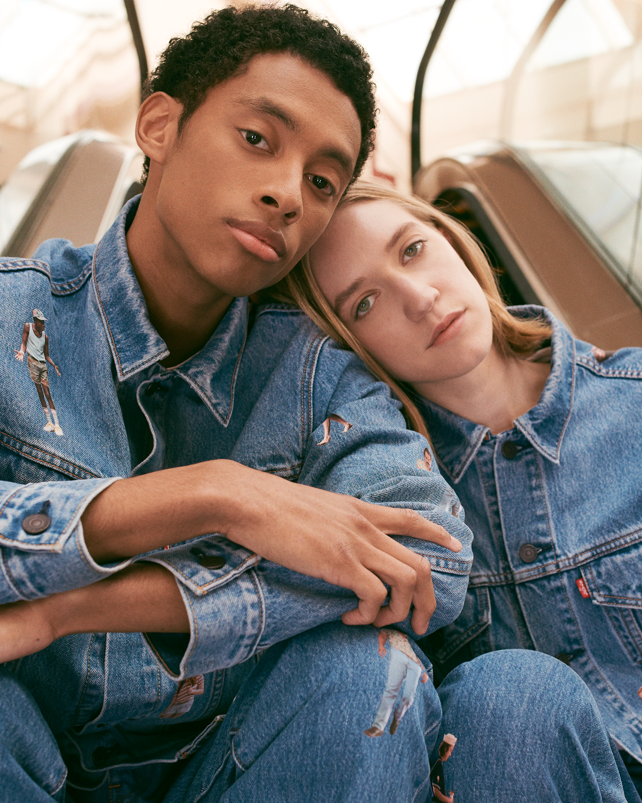 A young man and woman sitting closely together, wearing denim jackets, with the woman resting her head on the man's shoulder inside a building with high ceilings and large windows.