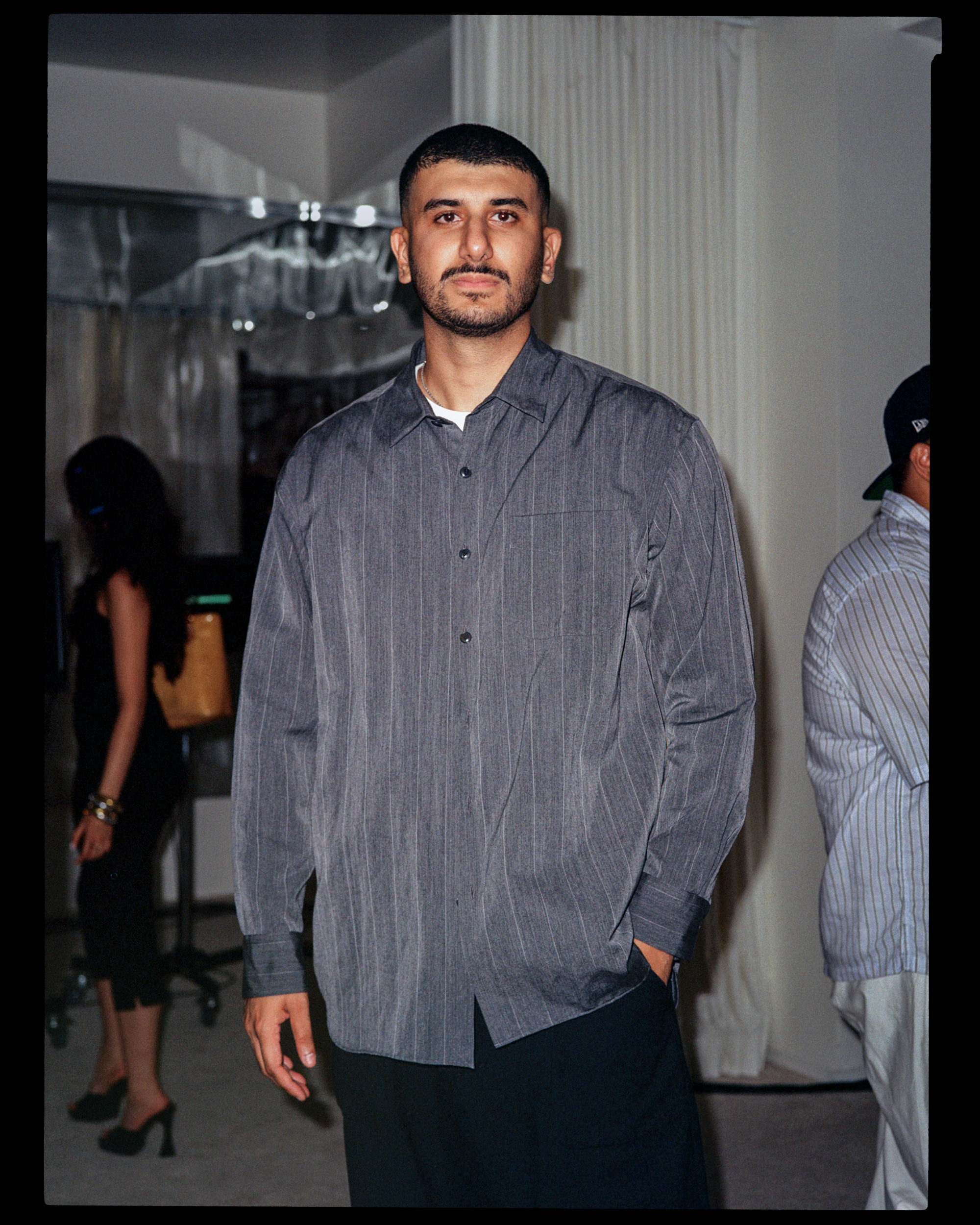 A man with short dark hair, beard, wearing a gray button-up shirt and dark pants, standing indoors with other people in the background.