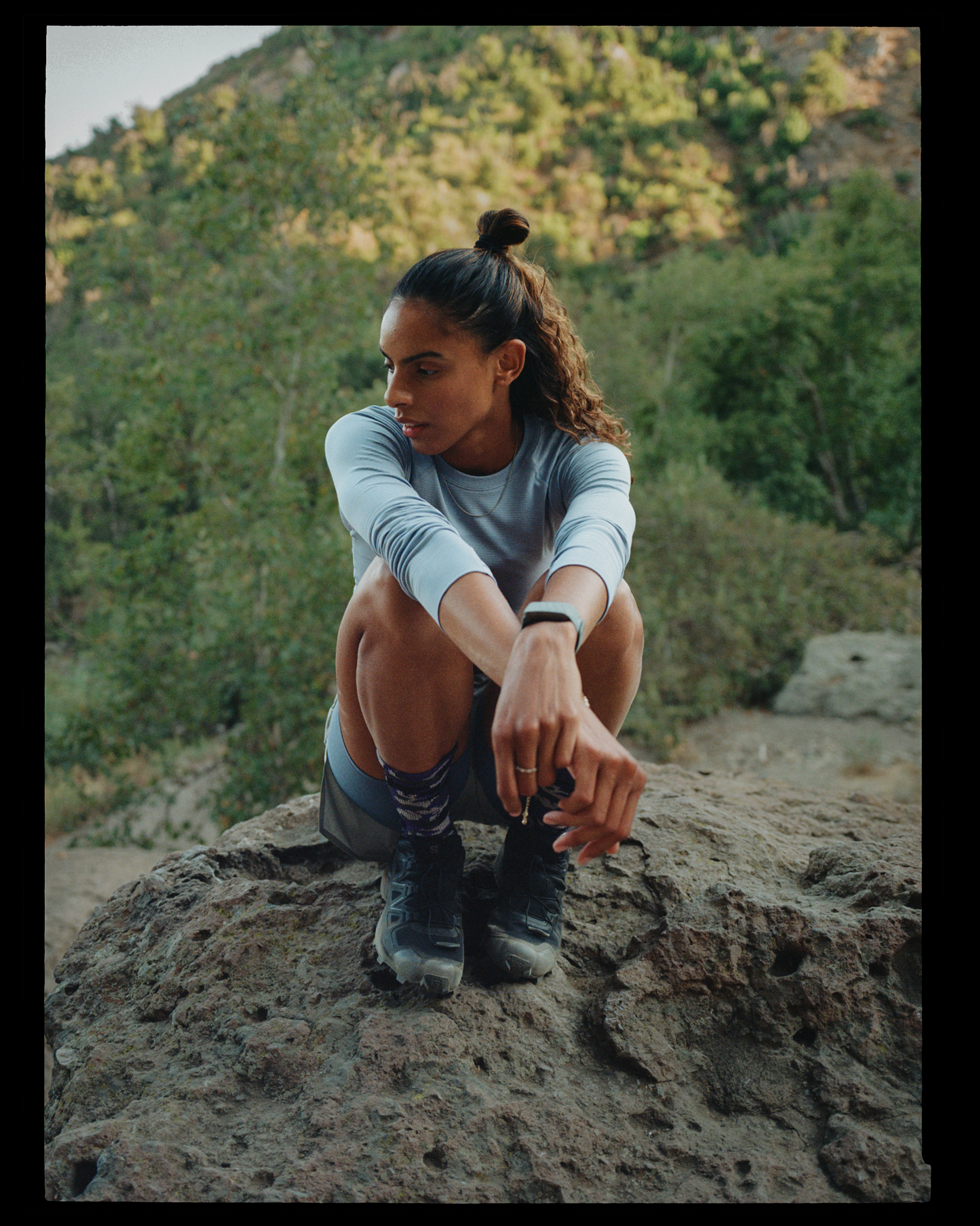A woman with curly hair tied up in a bun, wearing athletic clothing and sneakers, crouching on a large rock outdoors surrounded by trees and greenery.