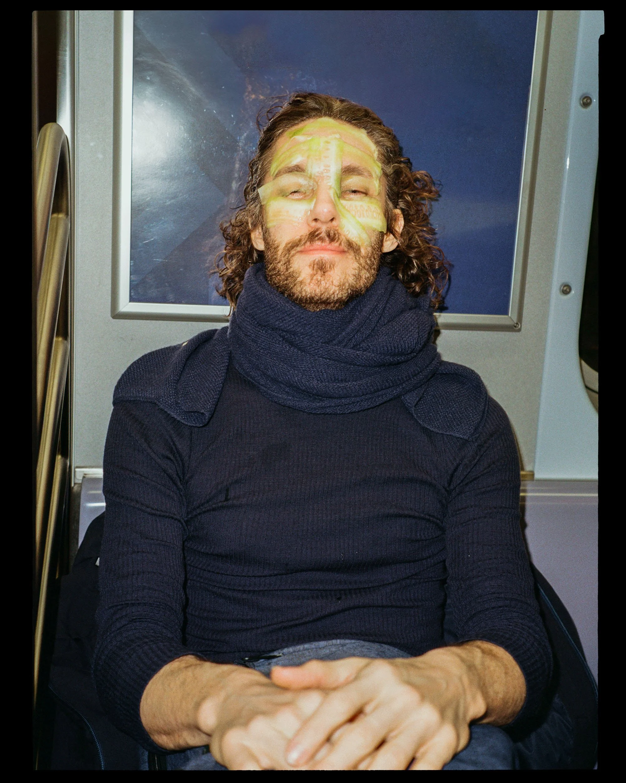 A man with curly hair and a beard wearing a dark sweater and scarf, lying back with eyes closed, covered with a yellow face mask, on a medical or spa table with a clear panel behind him.
