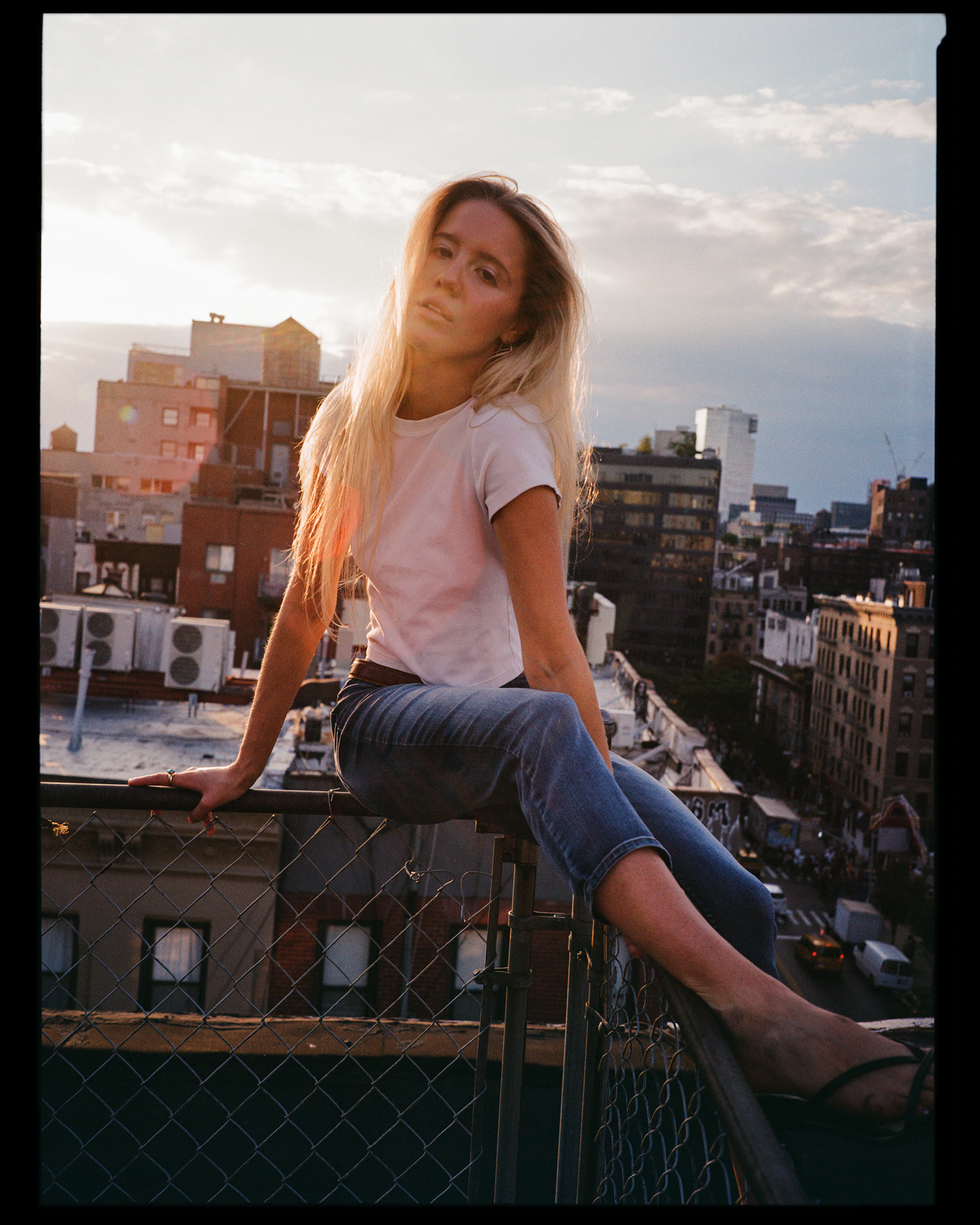 A young woman with long blonde hair wearing a white t-shirt and blue jeans sitting on a rooftop fence with an urban cityscape background during sunset.