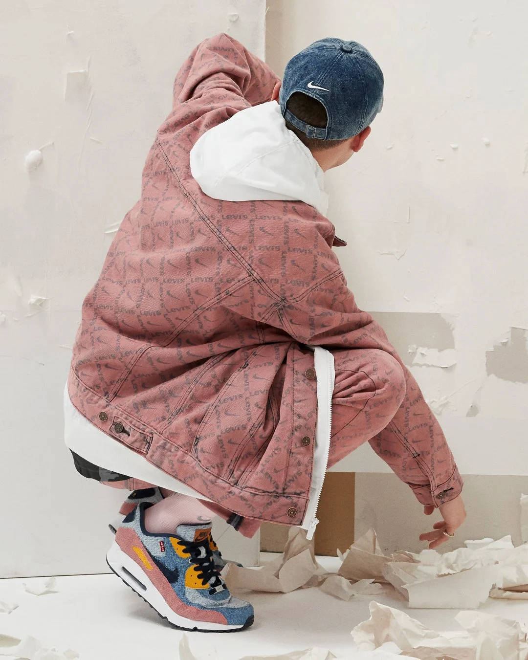 A person crouches down on a white floor, picking up crumpled brown paper, wearing a pink Levi's jacket, a white hoodie, a blue Nike cap, and colorful Nike sneakers.