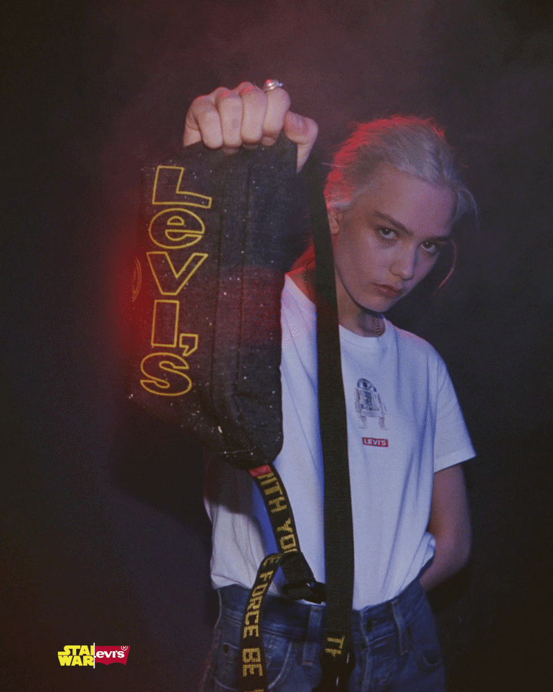 A young woman with blonde hair in a ponytail, wearing a white Levi's t-shirt, holding a black strap with yellow text. She has a serious expression and is standing against a dark background with red lighting. The strap has the words "SHOOT, HIDE, BE" and the person appears to be posing with a dark bag with yellow, red, and white text, and a small "Star Wars" and Levi's logo in the bottom left corner.