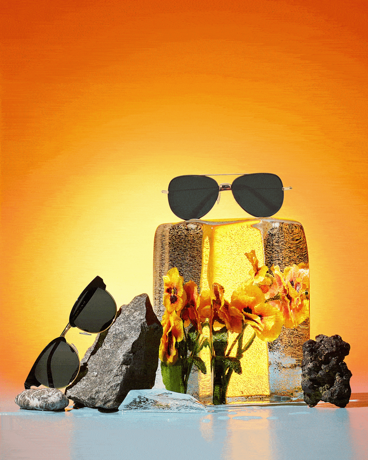 A centered pair of black sunglasses on a golden glass block, with flowers and rocks surrounding it on a blue surface with an orange background.
