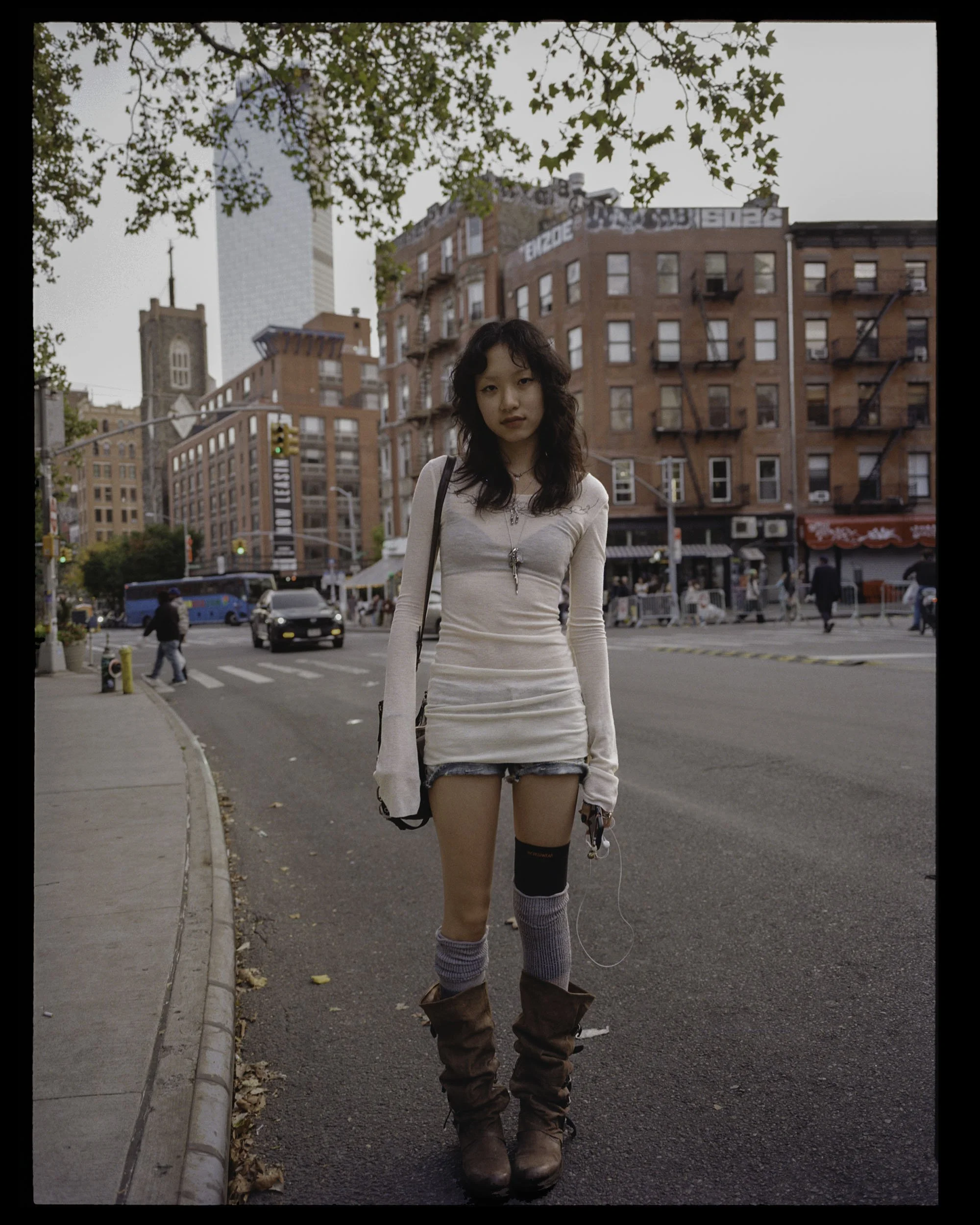 A young woman standing on a city street during daytime, wearing a white long-sleeve top, denim shorts, and brown boots, with a cityscape of brick buildings and trees in the background.
