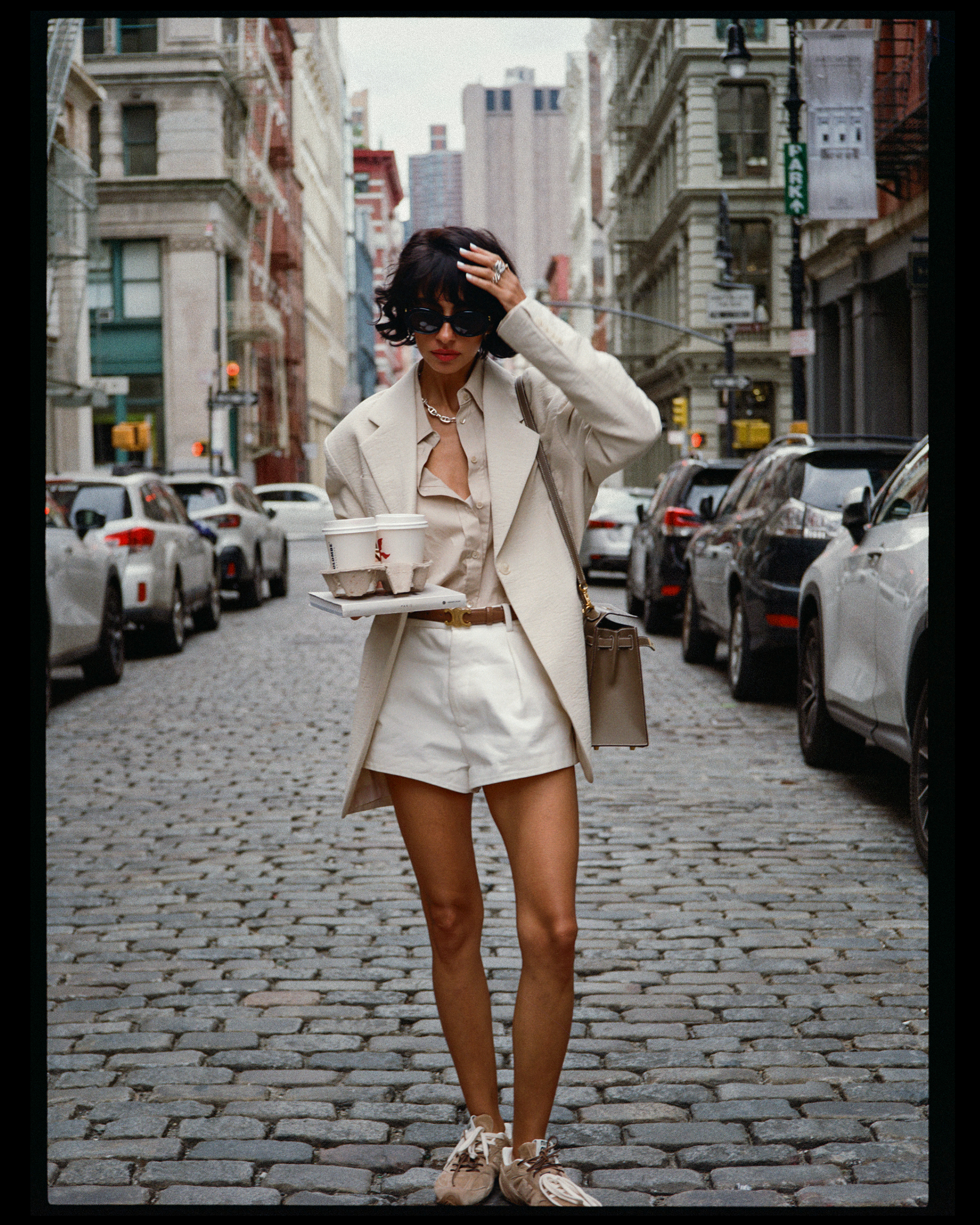 A woman with short dark hair wearing dark sunglasses, a beige blazer, matching shorts, and sneakers, walking on a cobblestone city street with parked cars and tall buildings.