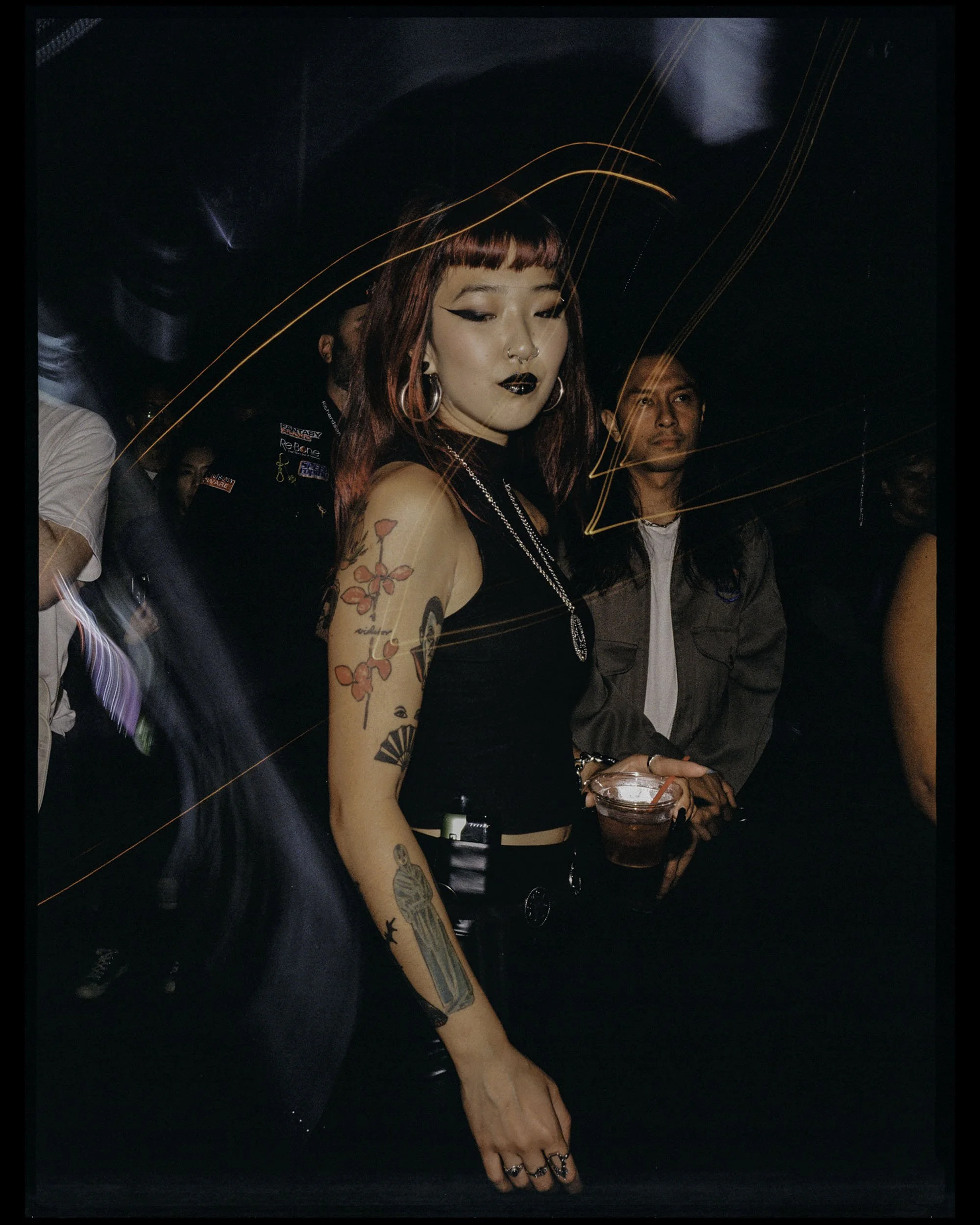 A woman with tattoos and dark makeup holding a drink at a party or nightclub, with light streaks and other people in the background.