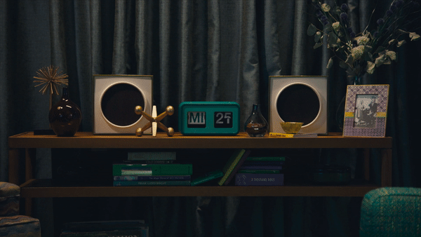 A wooden console table with decorative items including two square speakers, a gold abstract sculpture, a digital clock showing 25 minutes past 2, a brown vase, a small yellow dish, and a framed photo, set against dark curtains with a flower arrangement on the right.