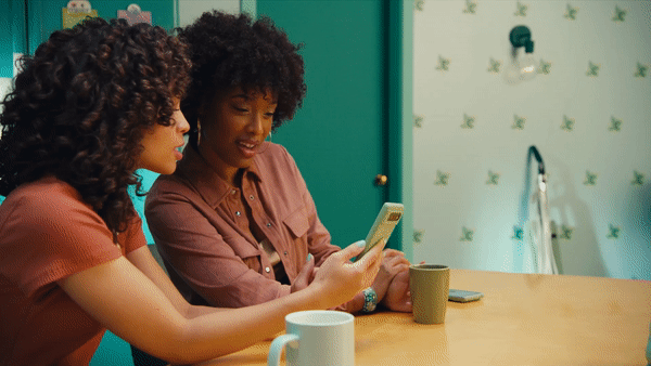 Two women sitting at a table, looking at a smartphone together in a kitchen with a green and white wall.