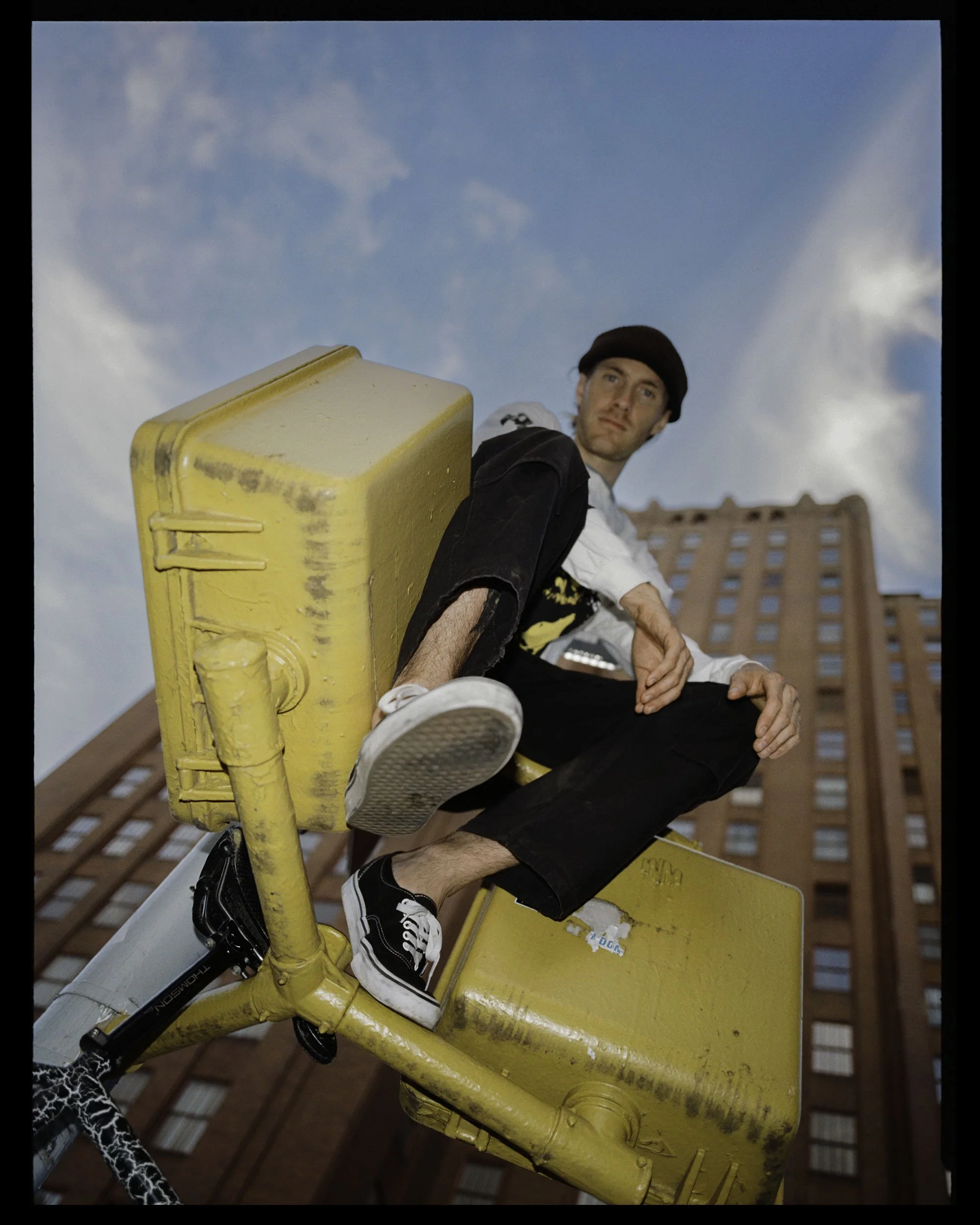 A young man with a beanie sitting on a yellow metal structure, looking into the distance, with a brick building in the background and a blue sky with clouds.